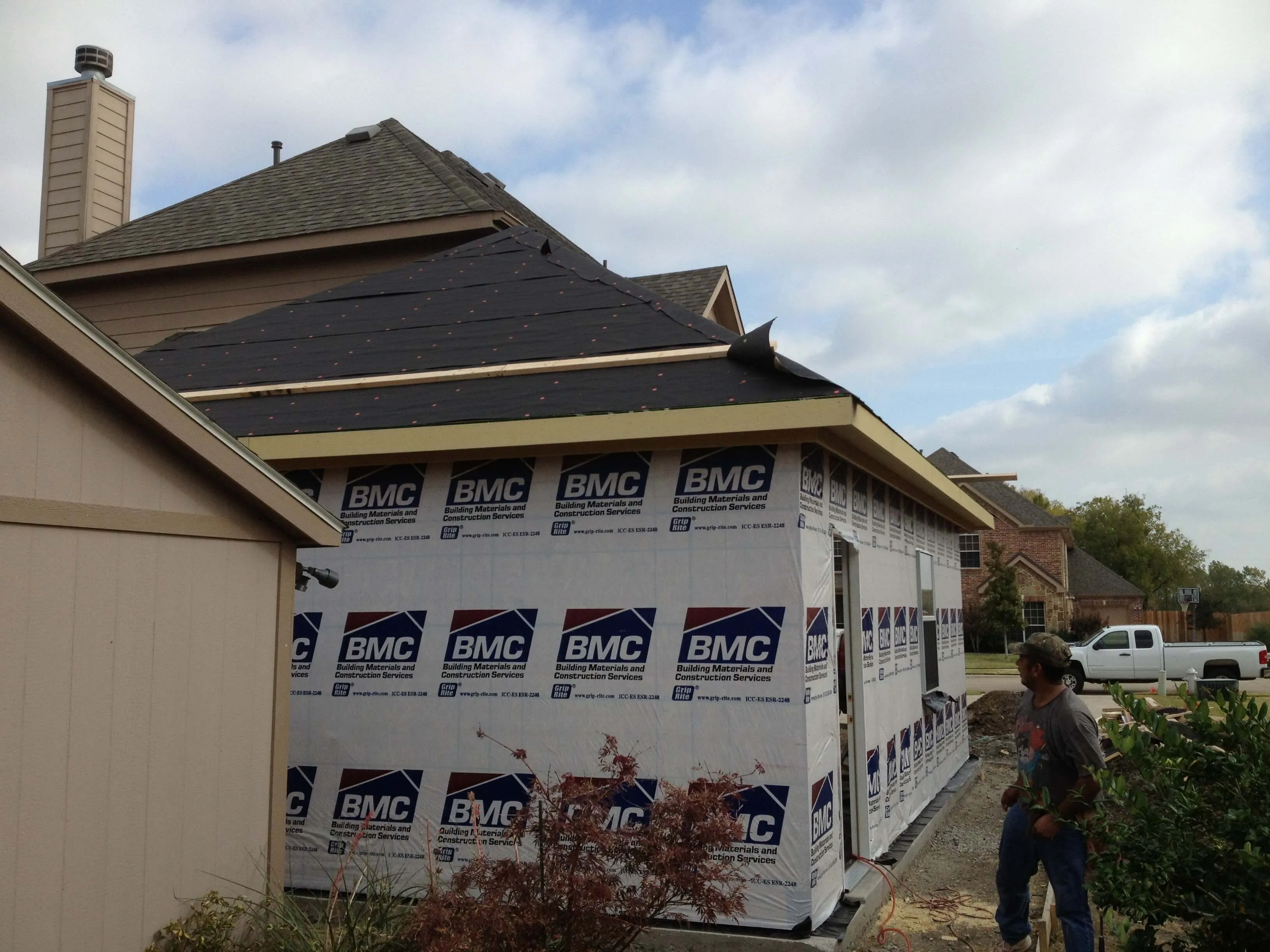 A partially constructed house wrapped with BMC building materials and a man standing near gravel driveway.