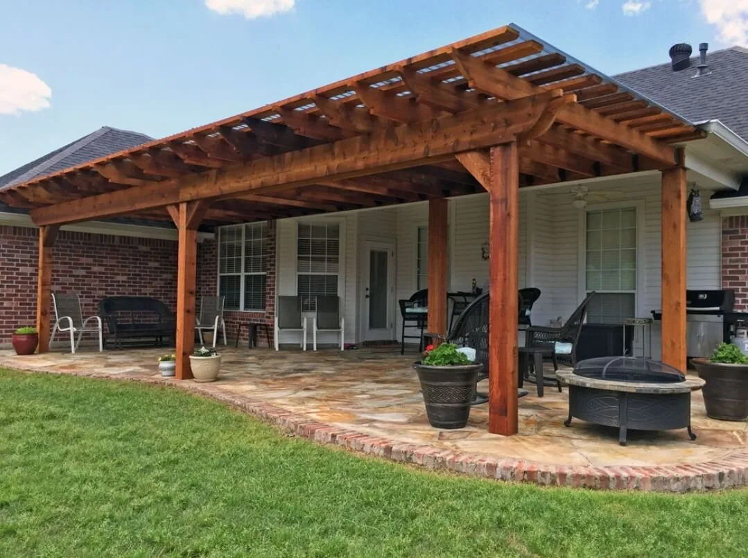 Covered patio with wooden pergola, stone floor, seating, fire pit, and potted plants in backyard.