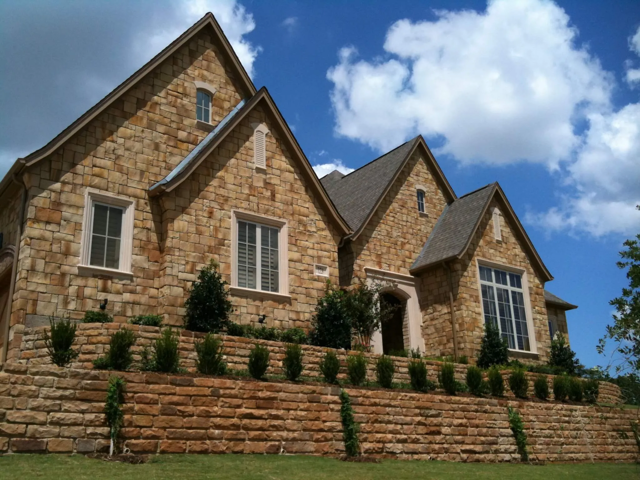 Large stone house with multiple gabled roofs and neatly trimmed bushes on terraced stone walls.