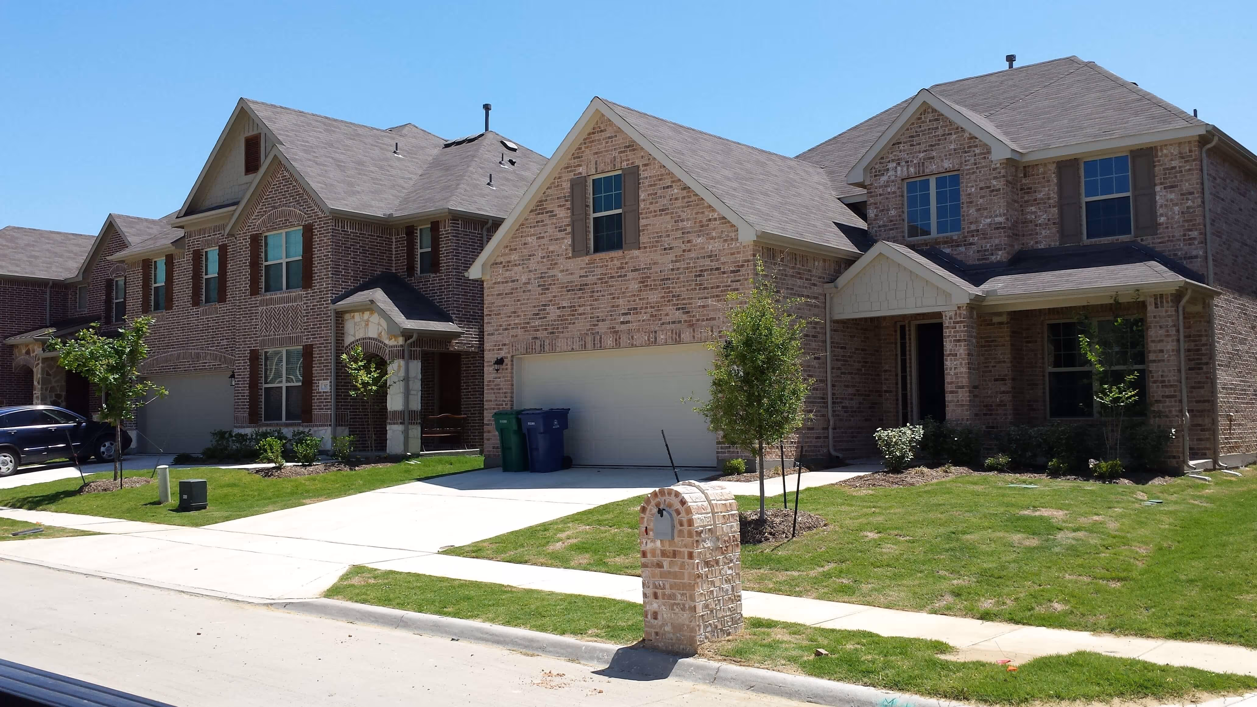 Suburban street view of two adjacent brick houses with garages, lawns, and a brick mailbox.