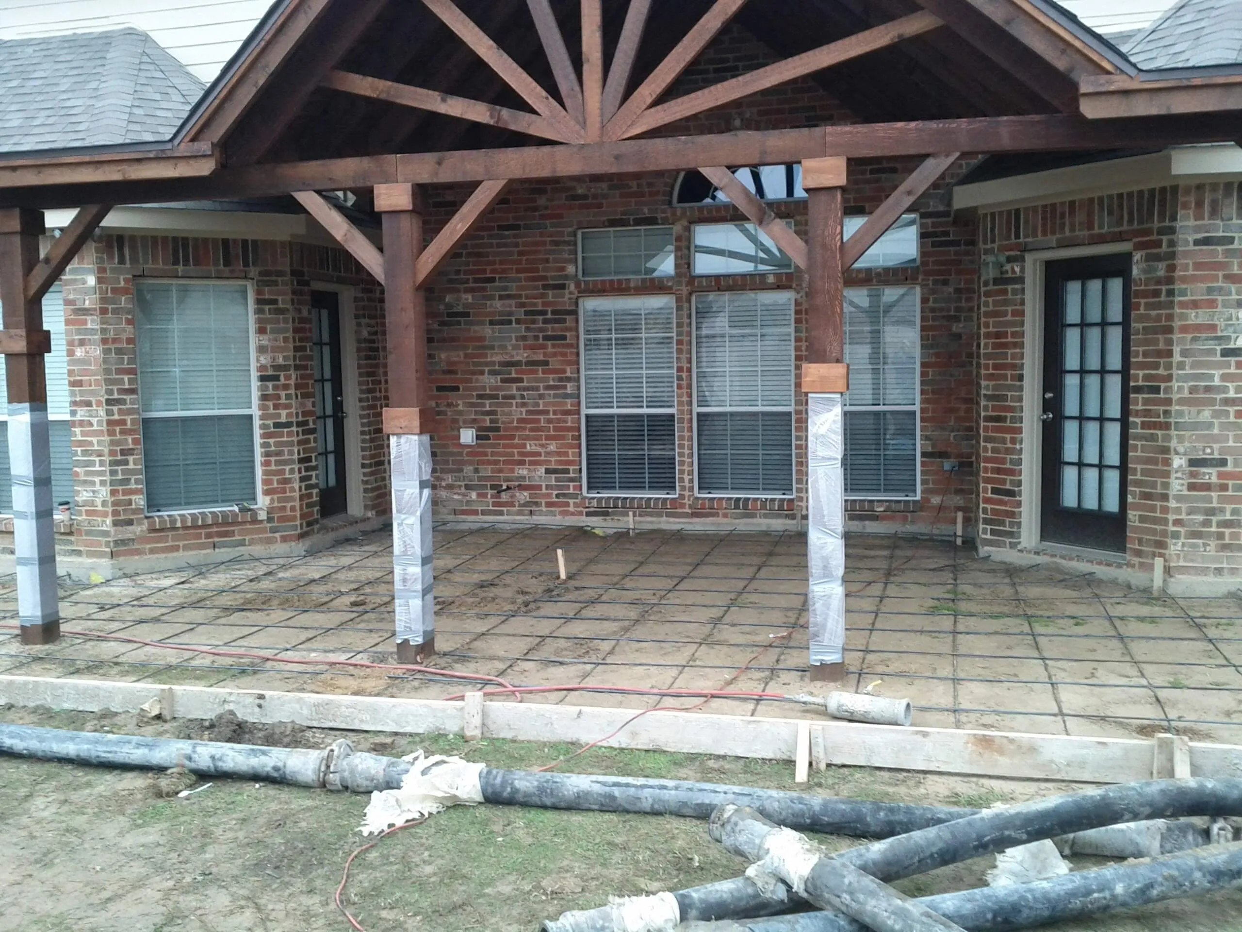 Back patio under construction with wooden framed roof, brick walls, and ground prepped for concrete pouring.