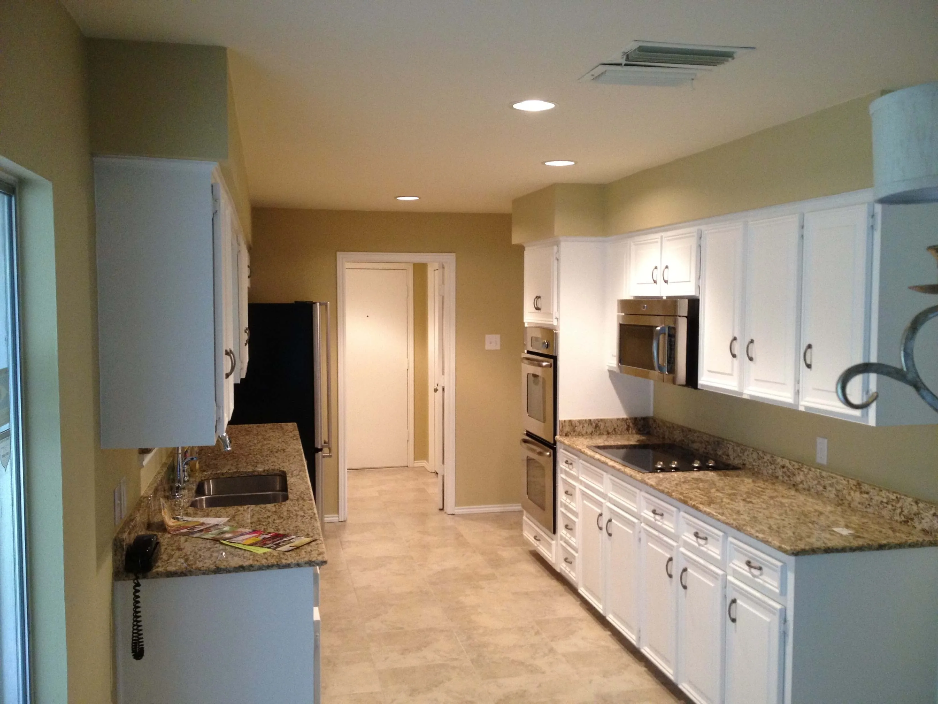 Narrow kitchen with white cabinets, granite countertops, double oven, stovetop, and a sink under a window.