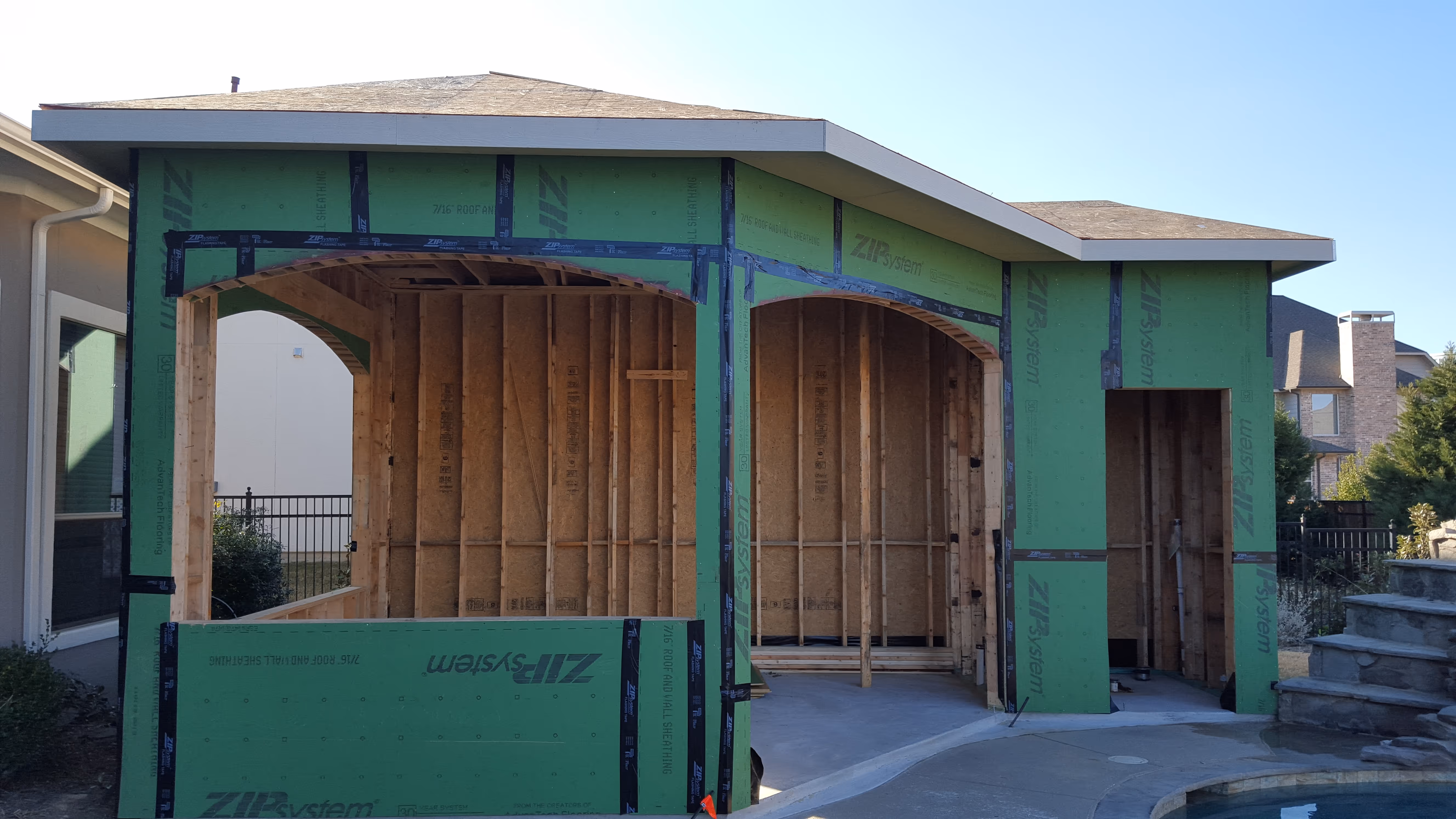Partially built green sheathed wooden structure with two large openings and one doorway beside a house.