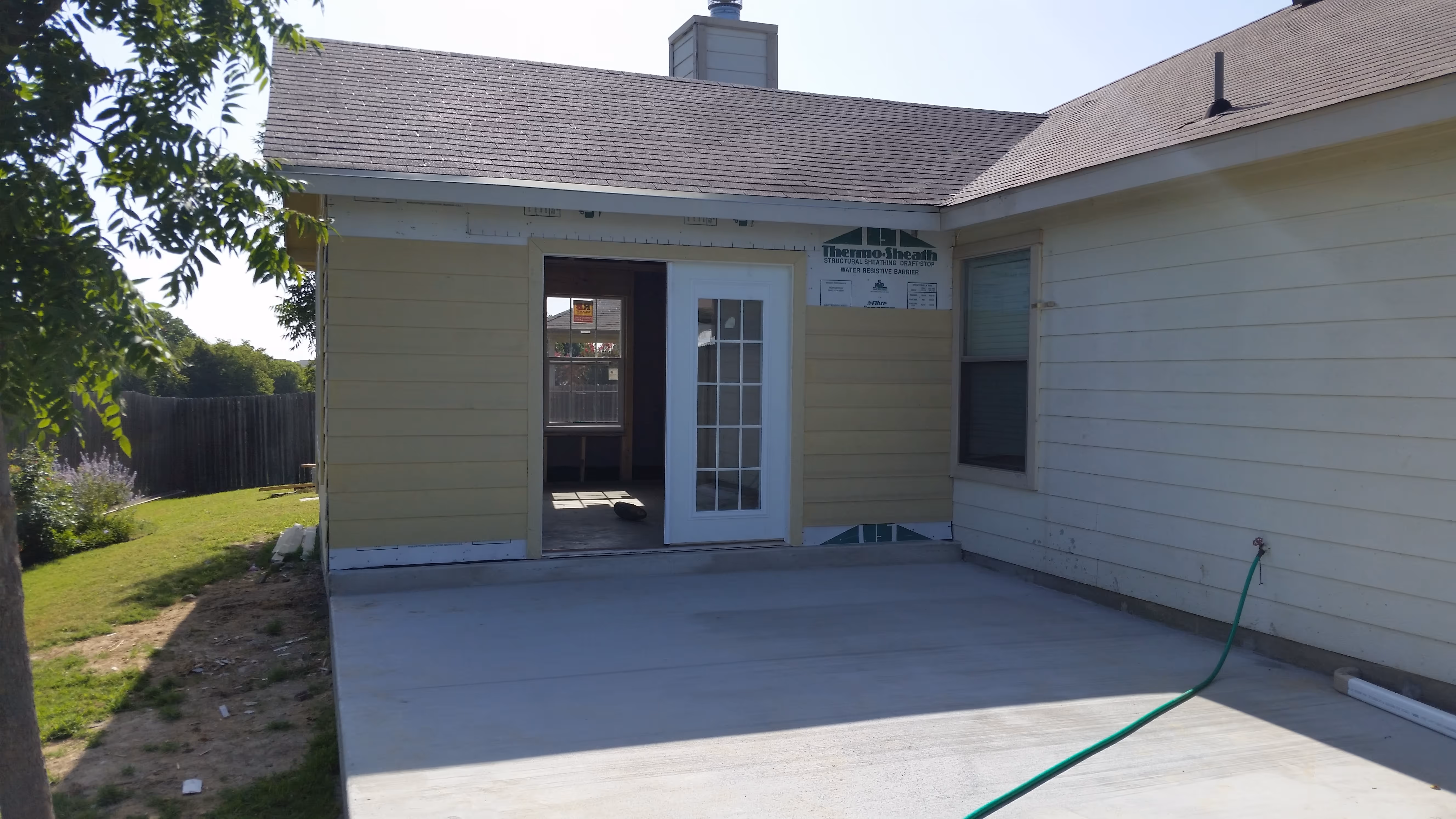 Backyard view of a house with beige siding, white door, window, and concrete patio with green hose.