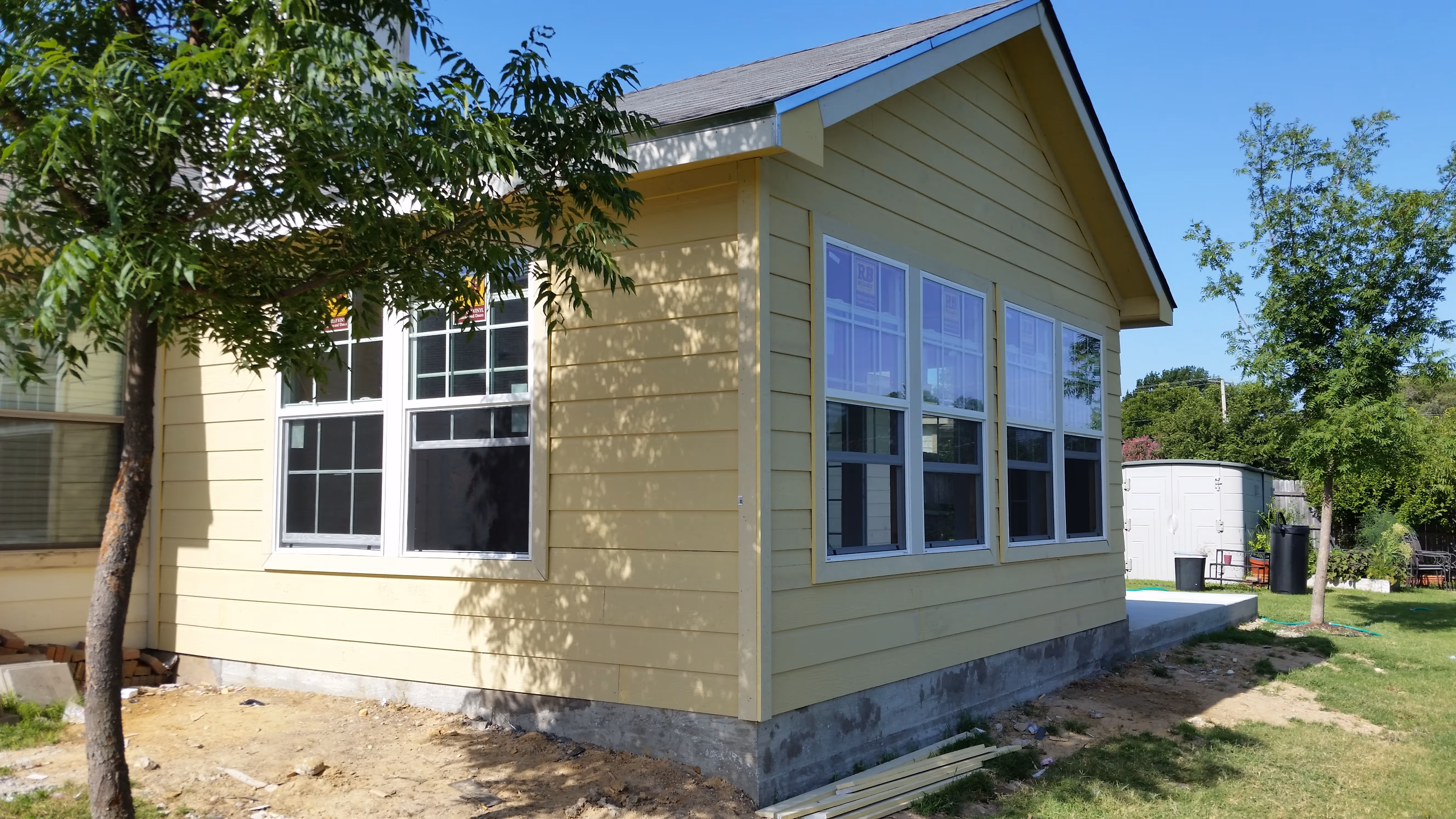 Yellow house corner with multiple windows, trees, and a white storage shed in the backyard.