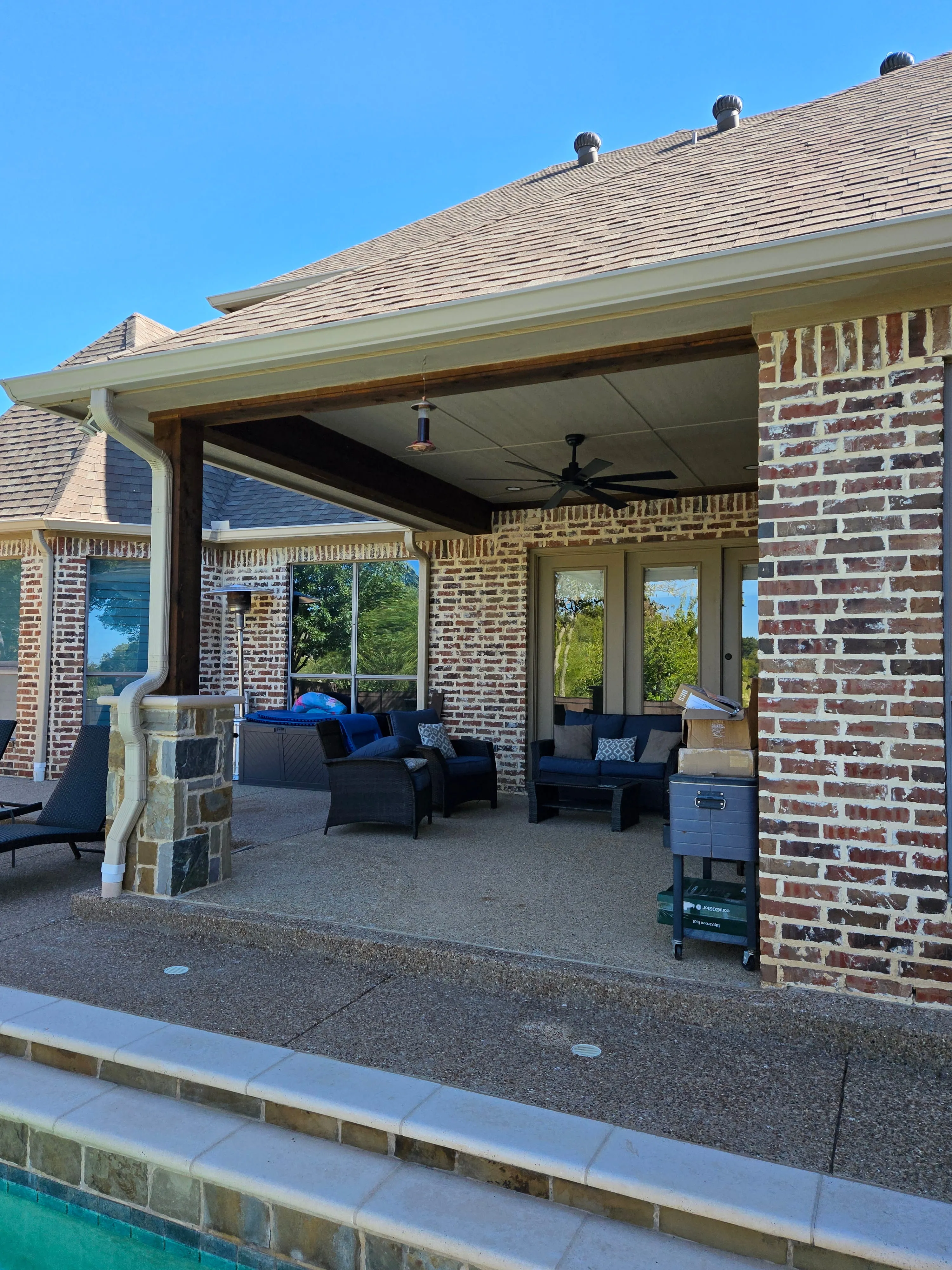 Covered patio with wicker furniture, ceiling fan, and poolside view under a clear blue sky.