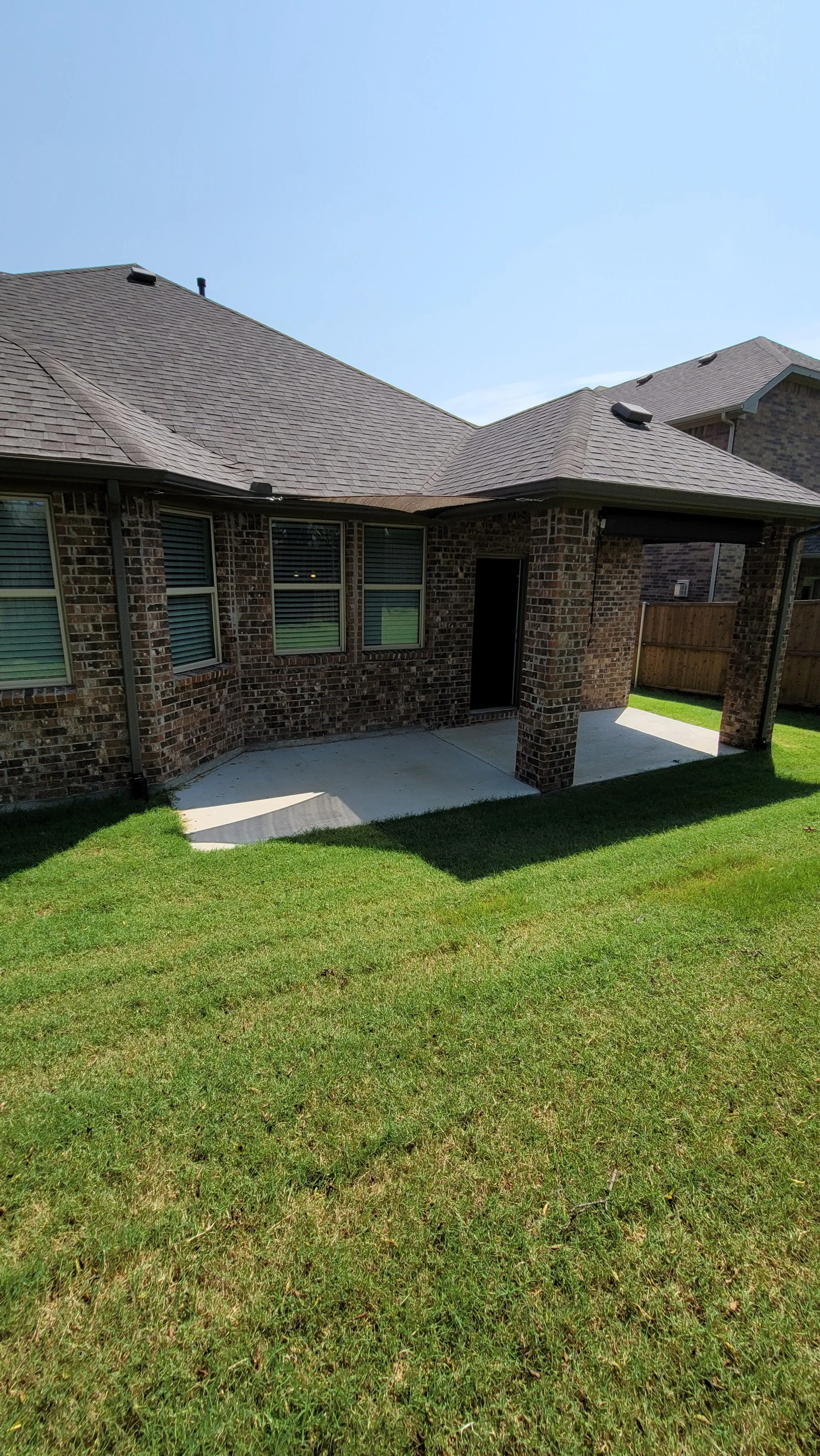 Backyard view of a brick house with covered concrete patio and green grass under clear sky.