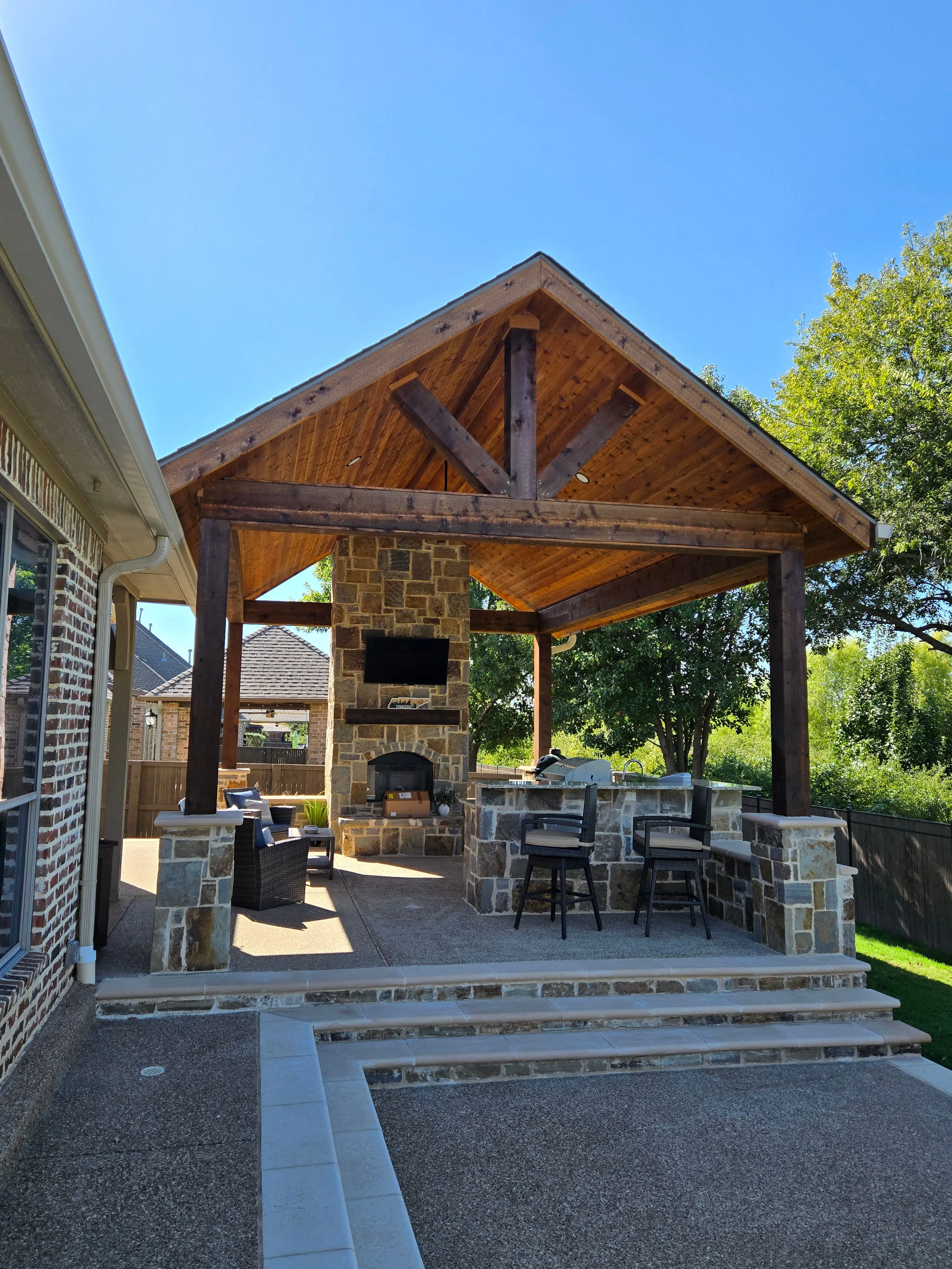 Outdoor covered patio with stone fireplace, bar counter, and seating under a wooden roof.
