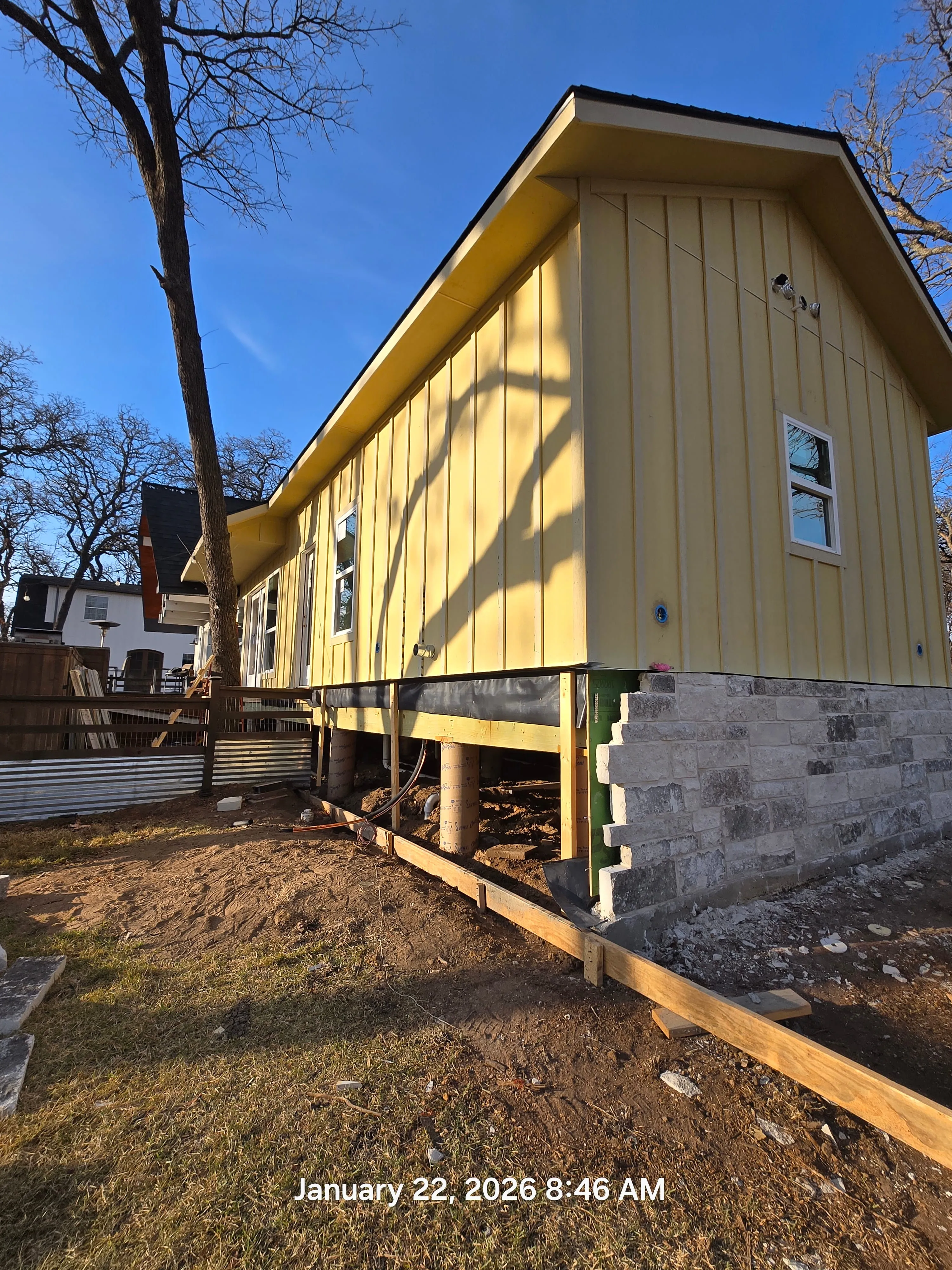 Yellow house elevated on concrete pillars with exposed foundation and shadows of tree branches.
