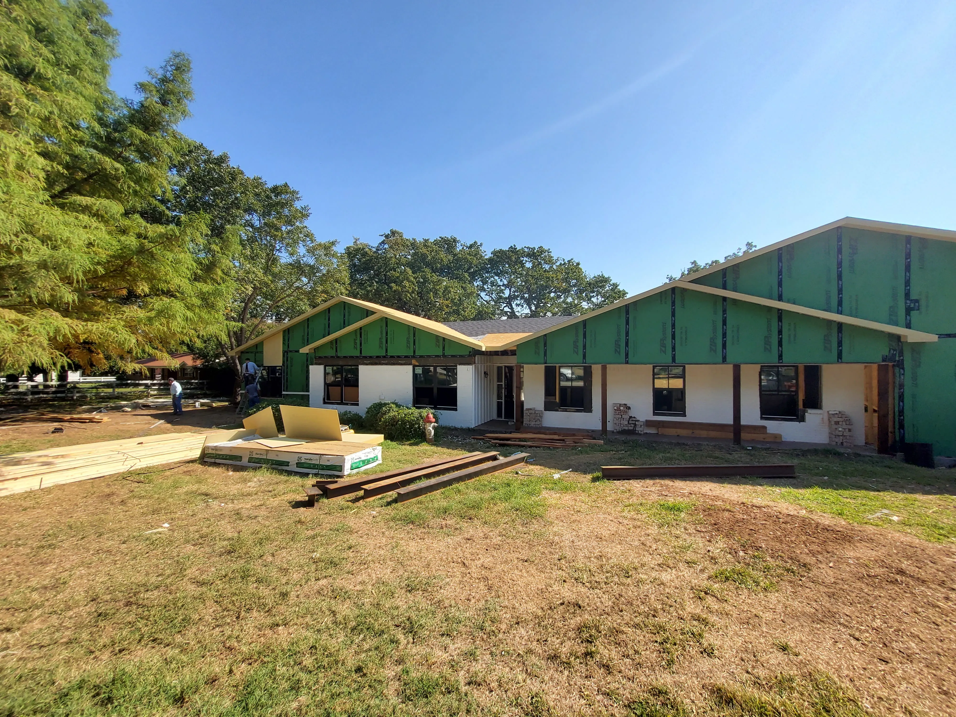 Partially constructed house with green sheathing, white brick walls, wooden beams, and construction materials on grass.