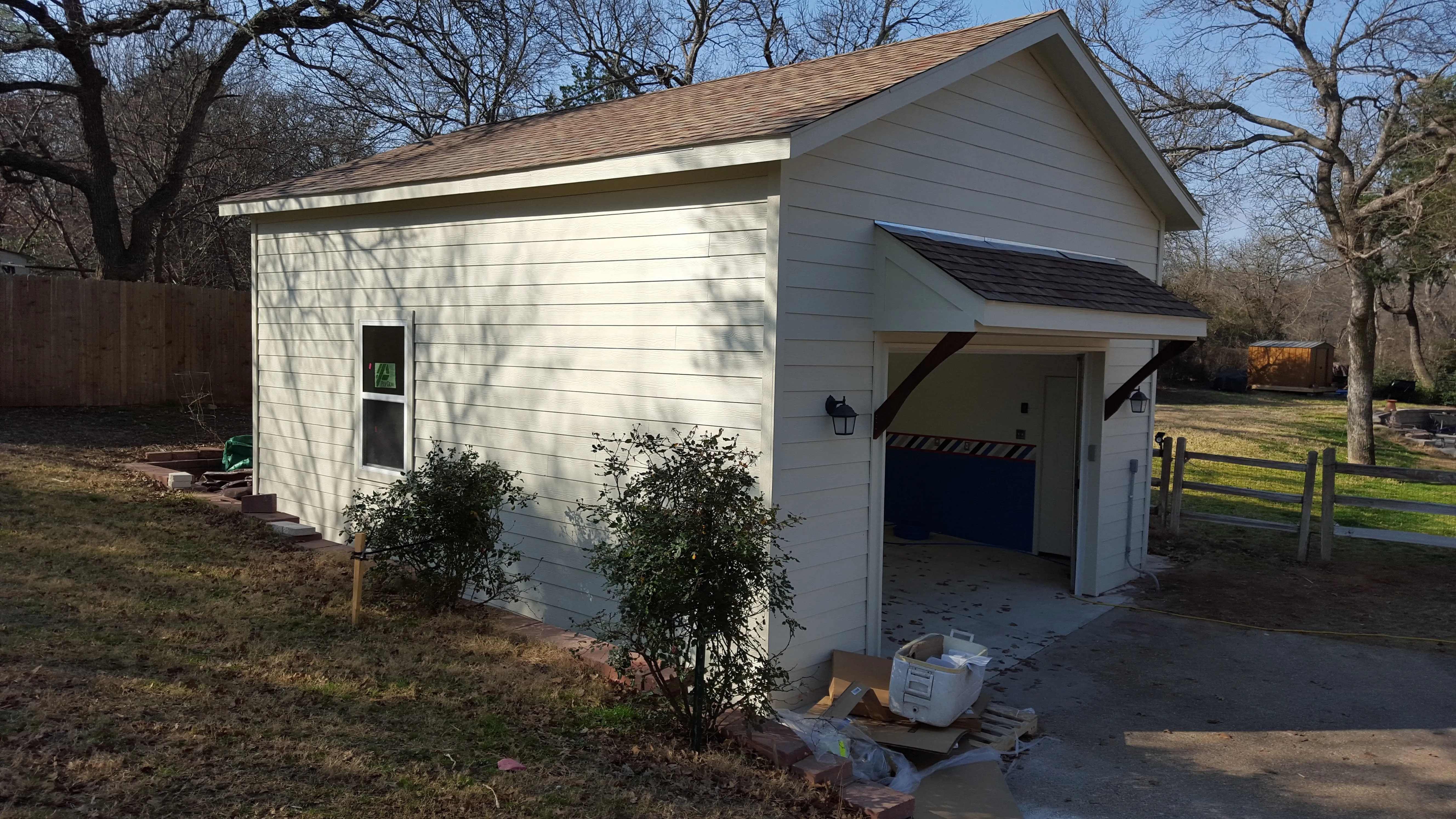 Light-colored garage with open door, bushes, and construction debris in a sunny yard with trees.