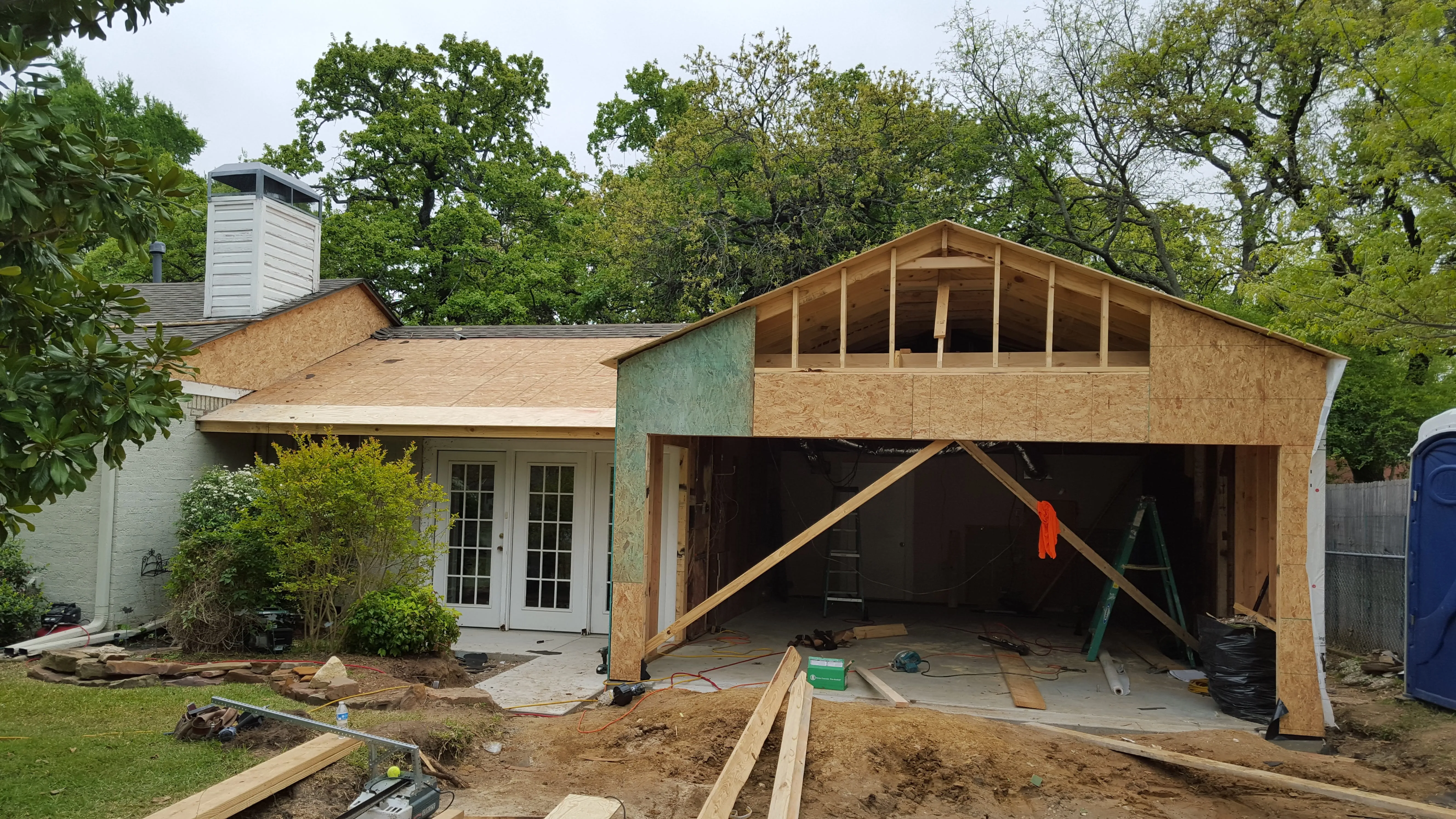 House under construction with wooden framework and open garage in a green, wooded yard.