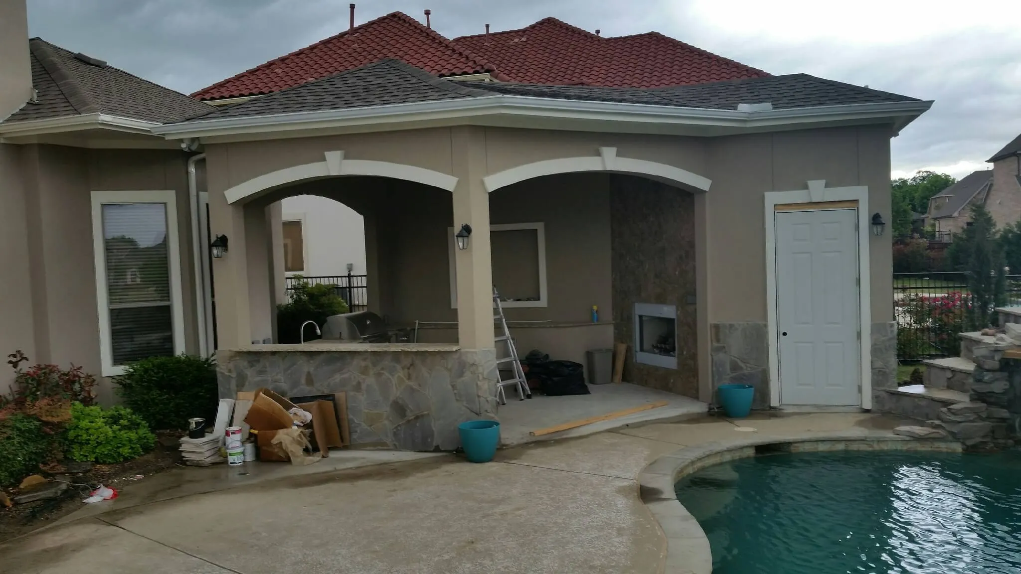 Backyard patio with stone bar, ladder, wood pile, and swimming pool under cloudy sky.