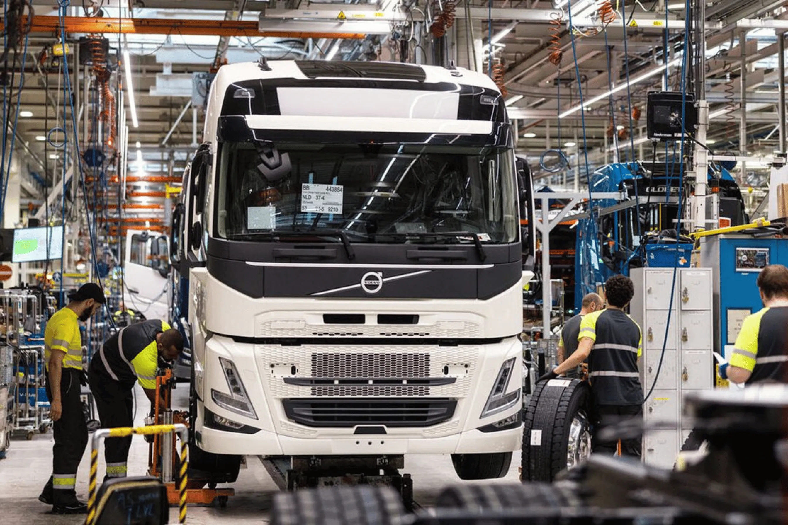 Workers assembling tires on large white Volvo truck in a busy industrial factory.