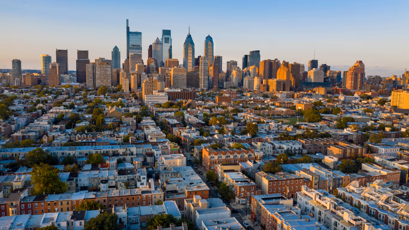Aerial view of a city with low residential buildings and a skyline of tall skyscrapers at sunset.