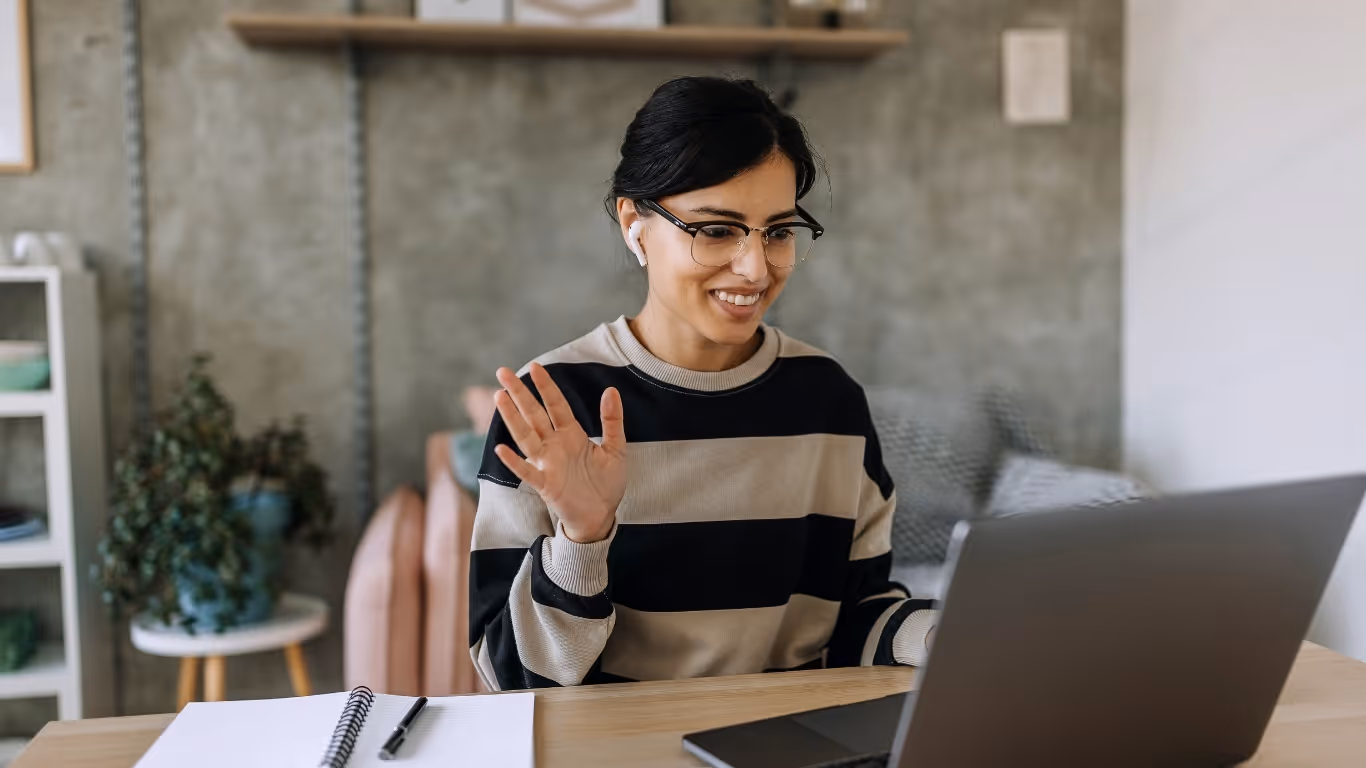 Smiling woman with glasses waving during a video call on her laptop while wearing earbuds.