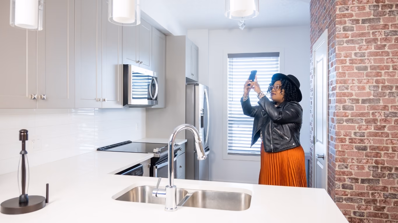 Woman wearing hat and leather jacket taking photos with smartphone in modern kitchen with brick wall.