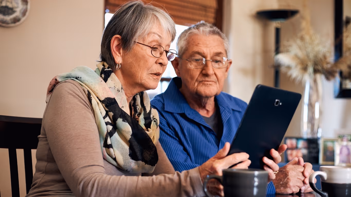 Older couple sitting and looking at a tablet together at a table with coffee mugs nearby.