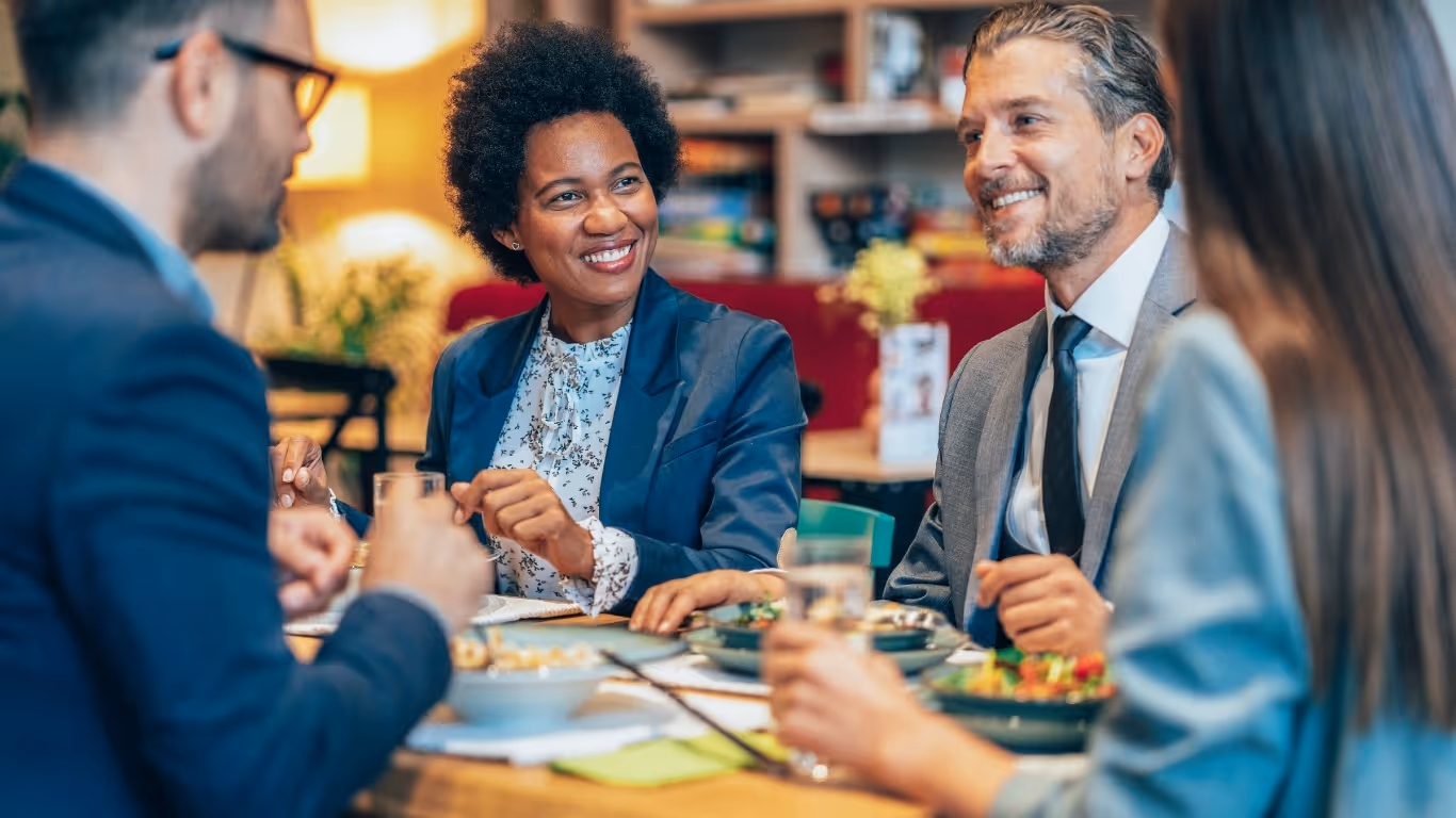 Four diverse business professionals smiling and chatting while having lunch in a casual office setting.