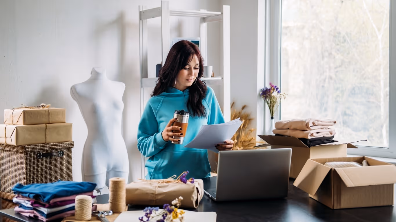 Woman in turquoise hoodie holding papers and coffee in a sewing studio with fabric and boxes.