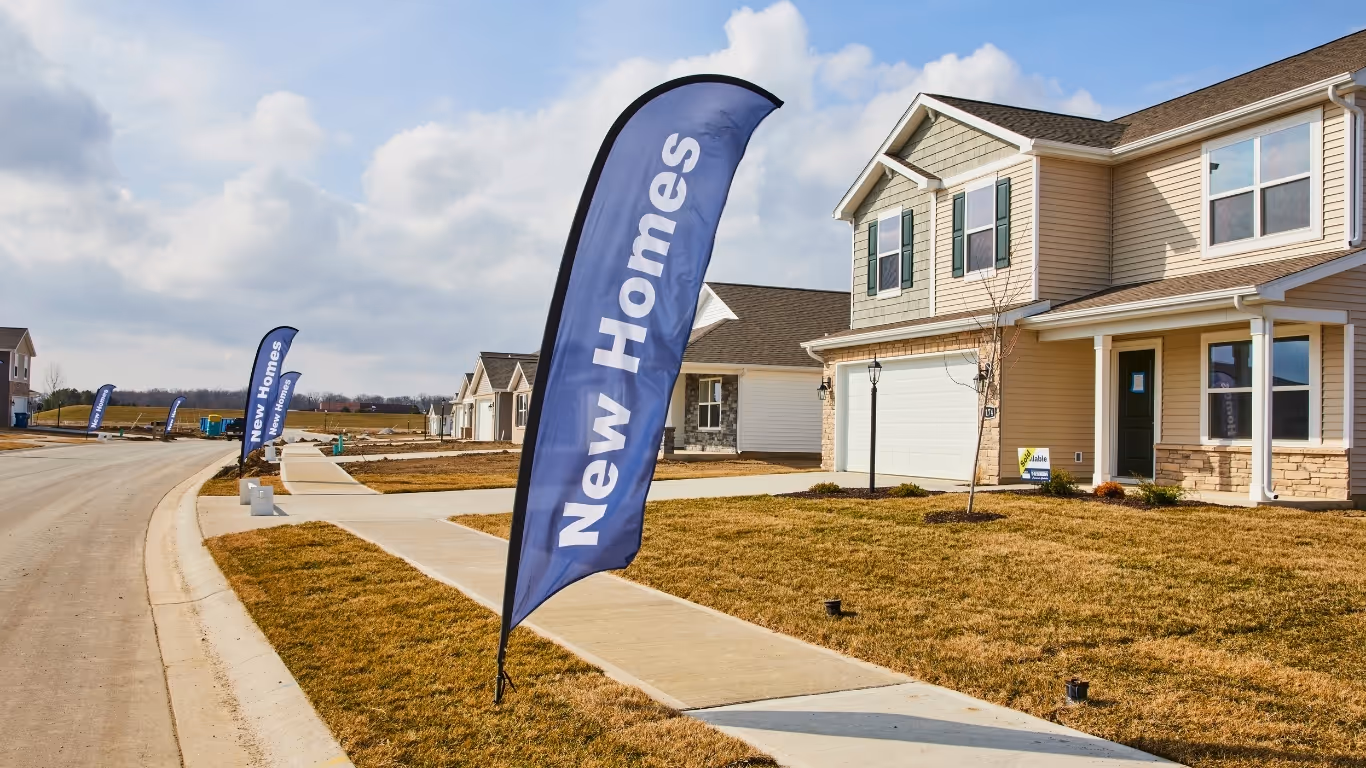 Suburban street with new homes for sale marked by blue flags on lawns under a partly cloudy sky.