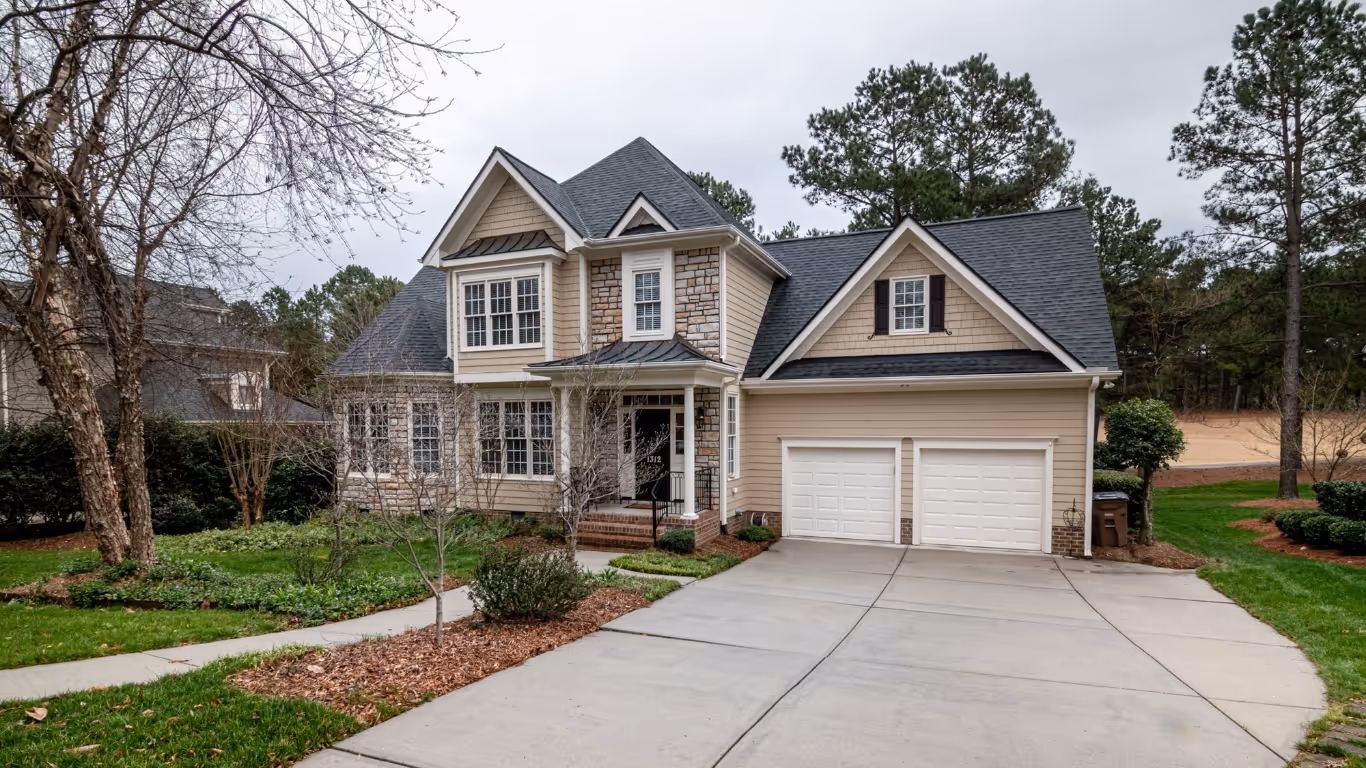 Two-story beige house with stone accents, a double garage, and a curved concrete driveway surrounded by greenery.