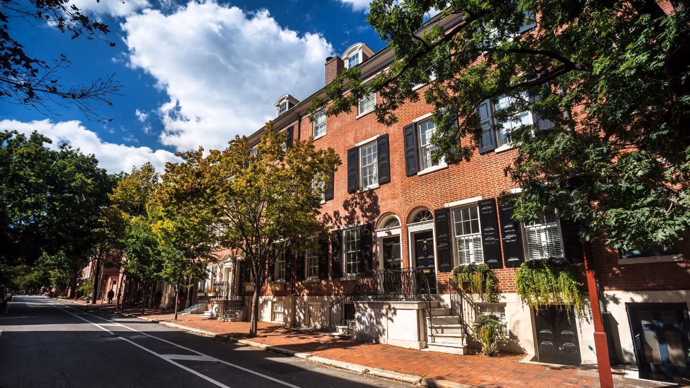 Row of red brick townhouses with black shutters and green trees on a sunny street.