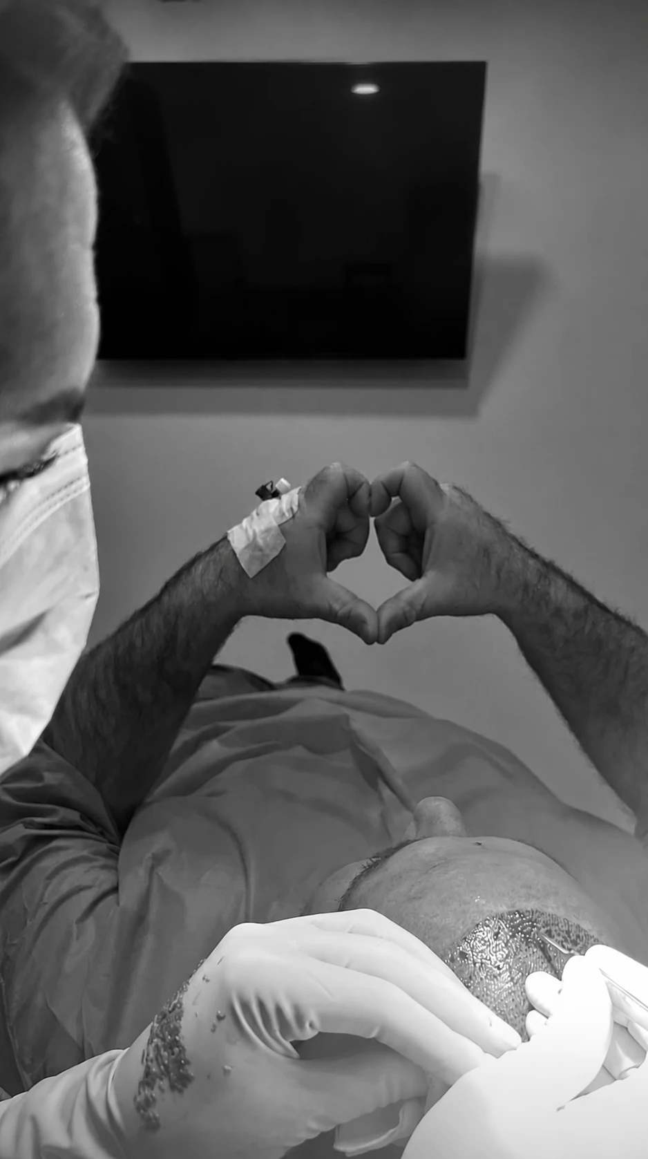 Patient mit Infusionskanüle formt ein Herz mit den Händen während medizinischer Behandlung am Kopf.