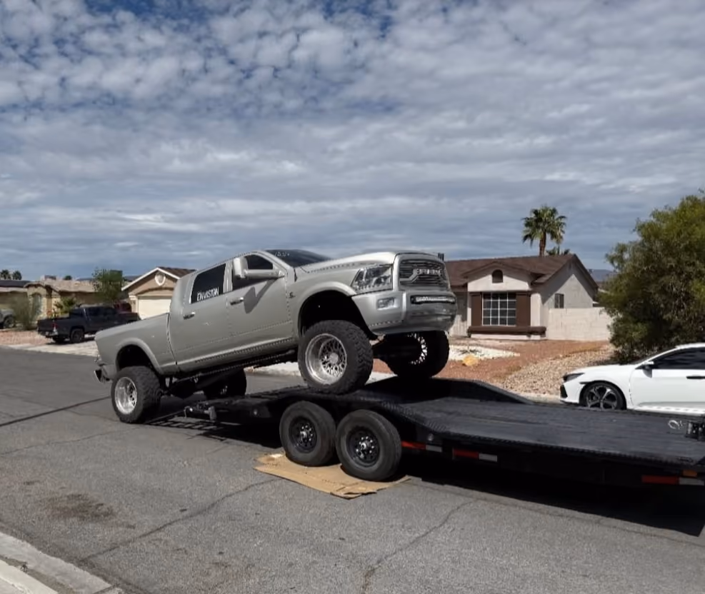 Silver lifted pickup truck driving onto a black car trailer on a residential street.