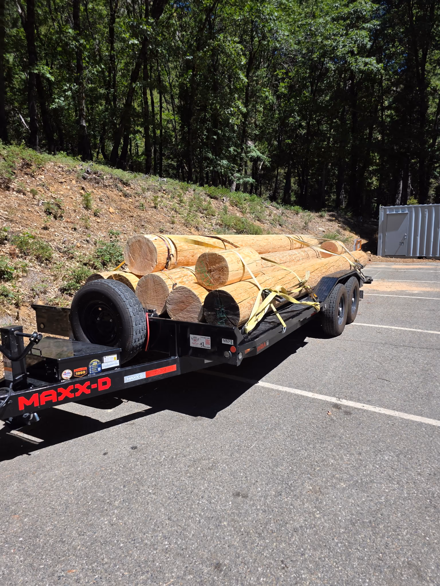 Black flatbed trailer hauling large round wooden logs secured with yellow straps in a forested parking area.