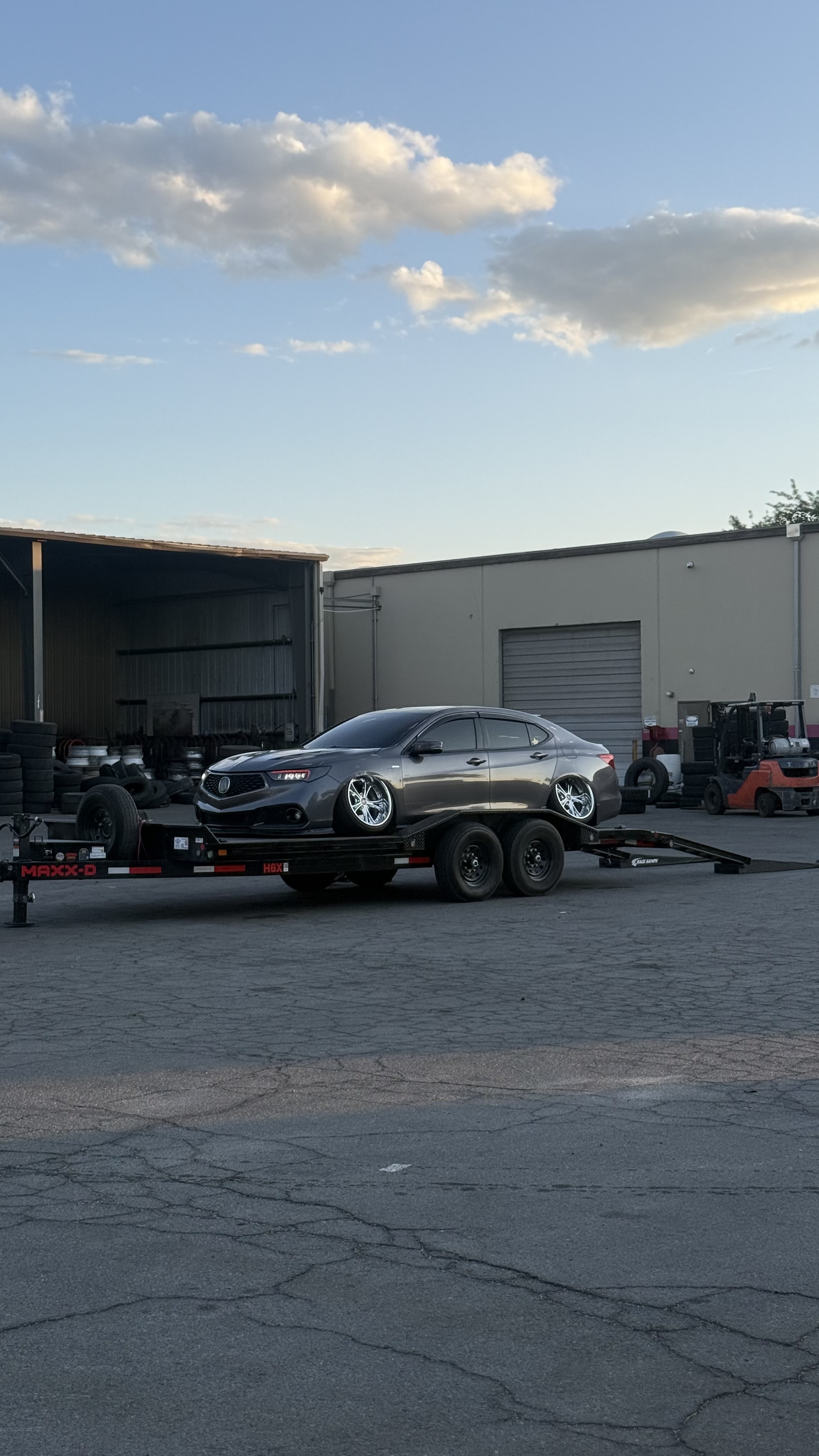 Black Audi sedan with heavily cambered wheels secured on a flatbed trailer in a parking lot with trees and hills in the background under a partly cloudy sky.
