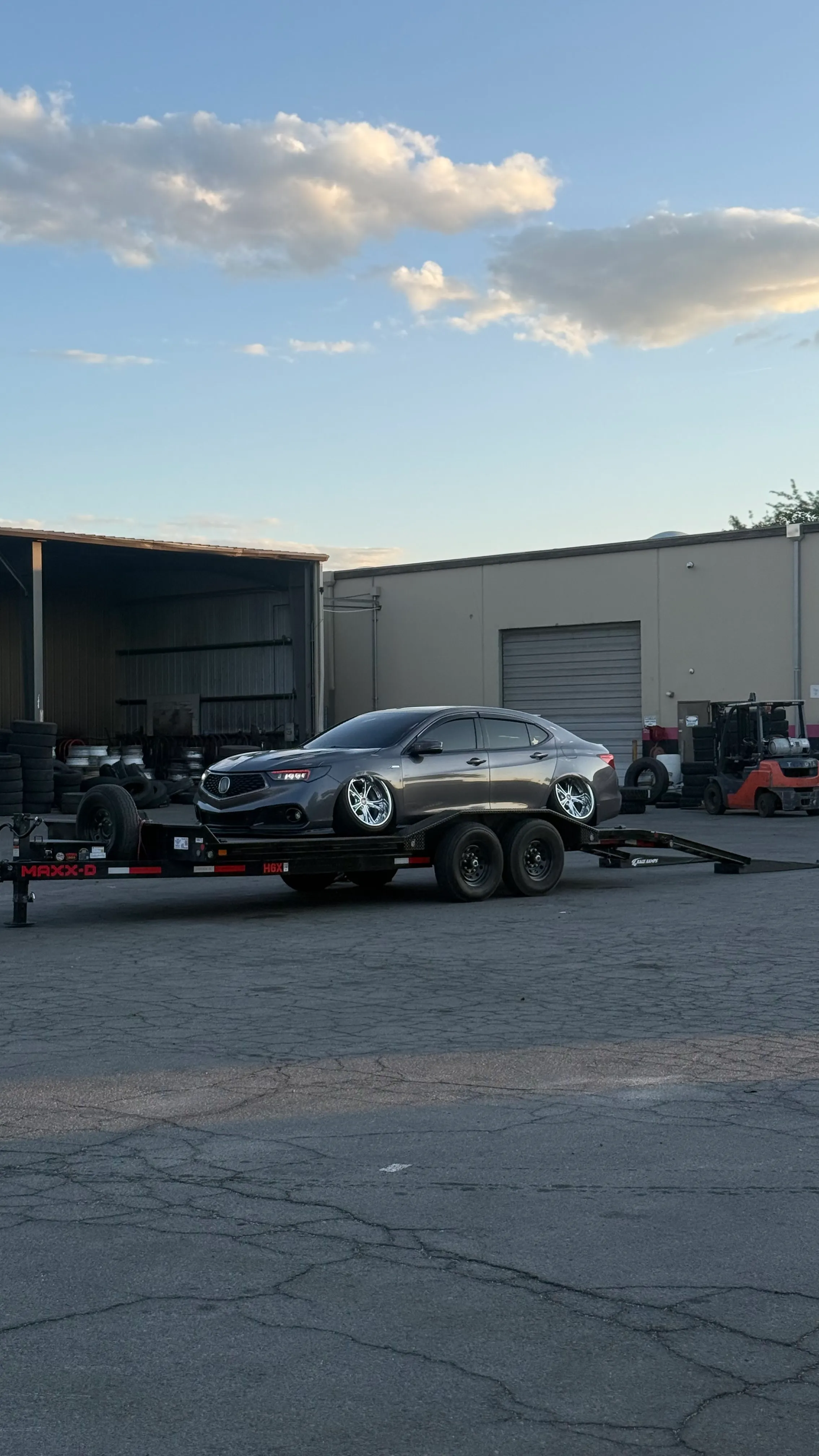 Grey sedan with shiny chrome rims on a flatbed trailer in an industrial area at dusk.