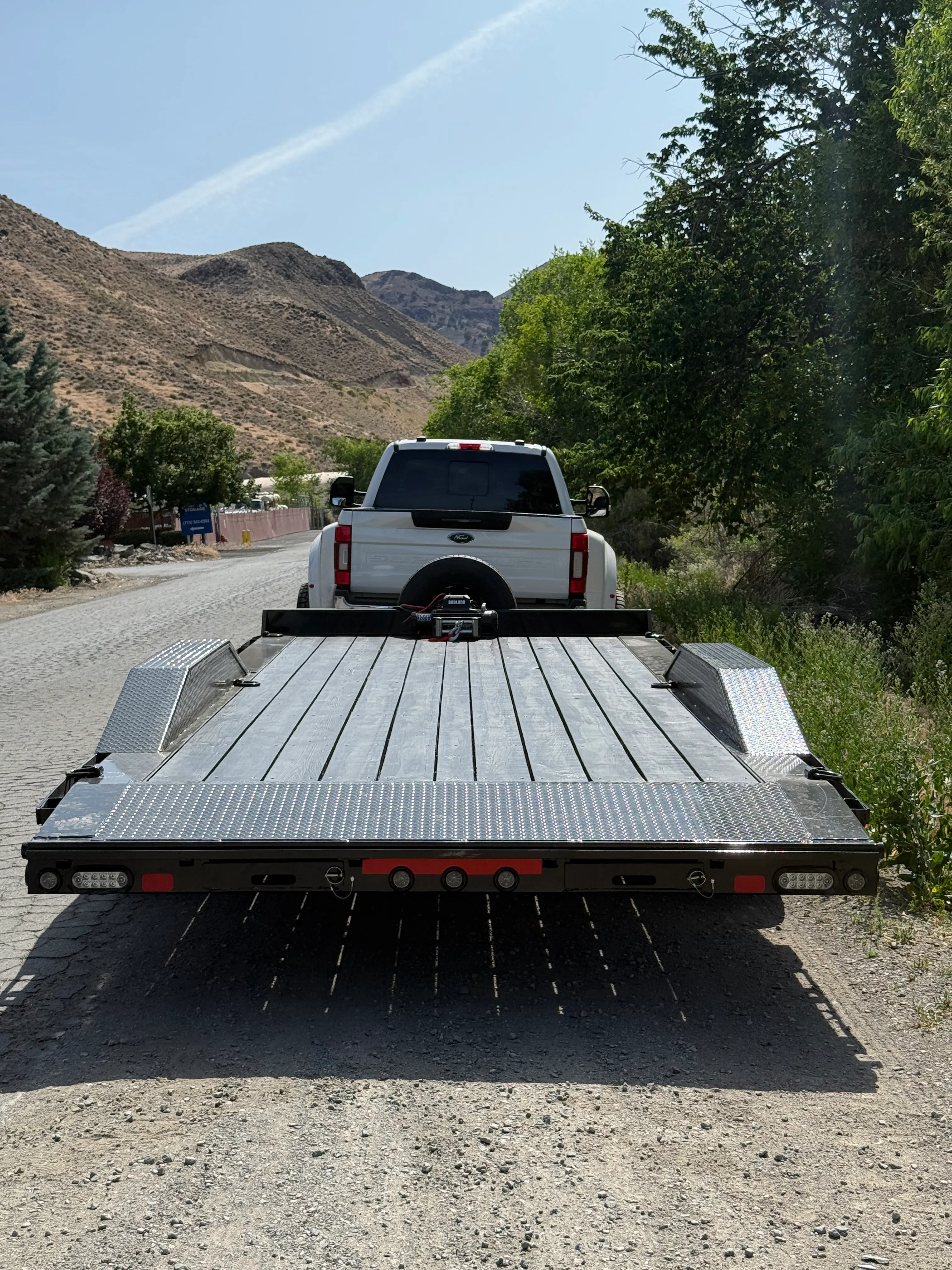 White pickup truck with empty flatbed car trailer on a gravel road, trees and hills in background.