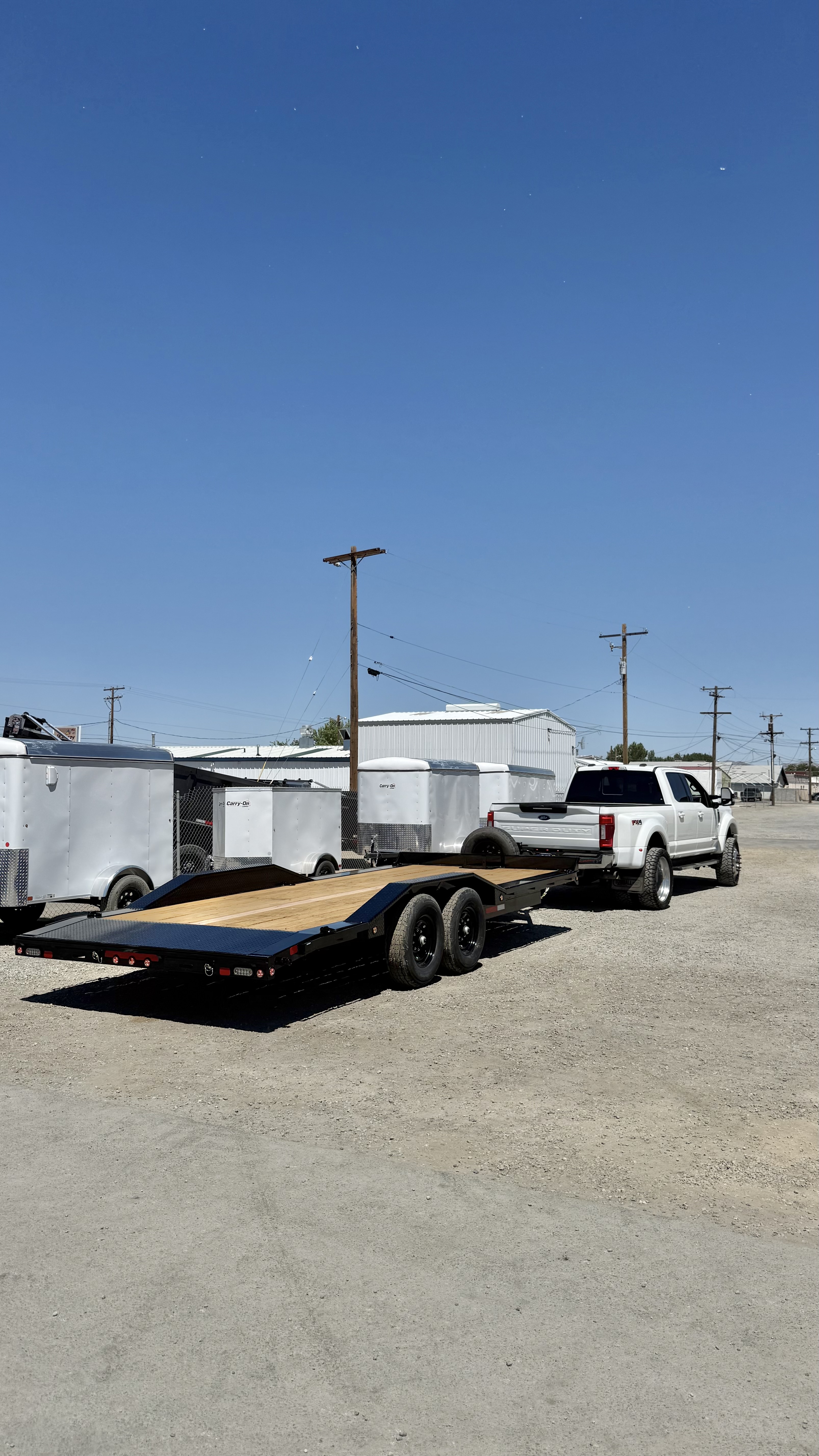 White pickup truck towing a black flatbed trailer with wooden floor, parked on gravel near white trailers and a metal building.