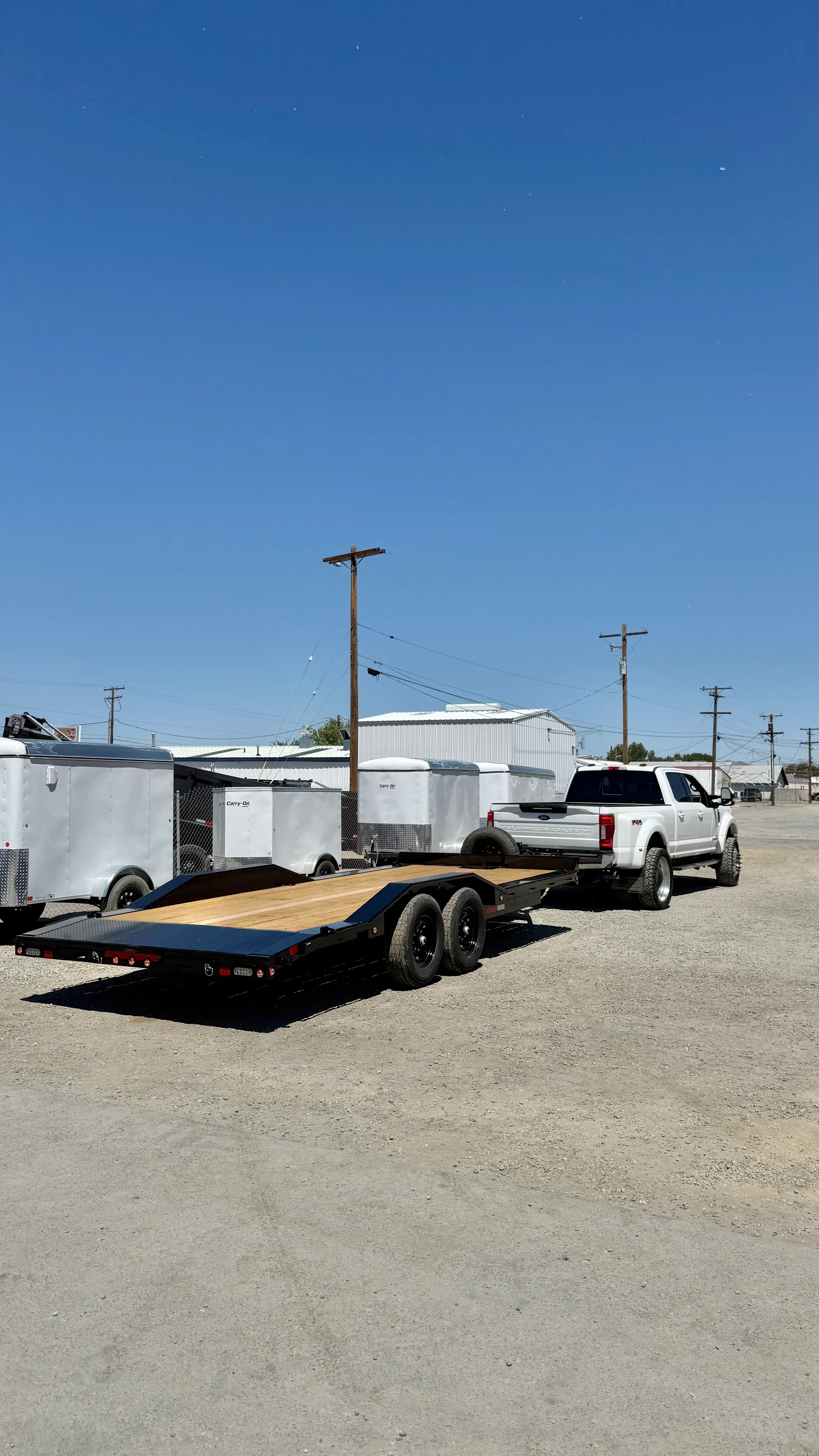 White pickup truck towing empty black flatbed trailer on a gravel lot with white trailers in background.