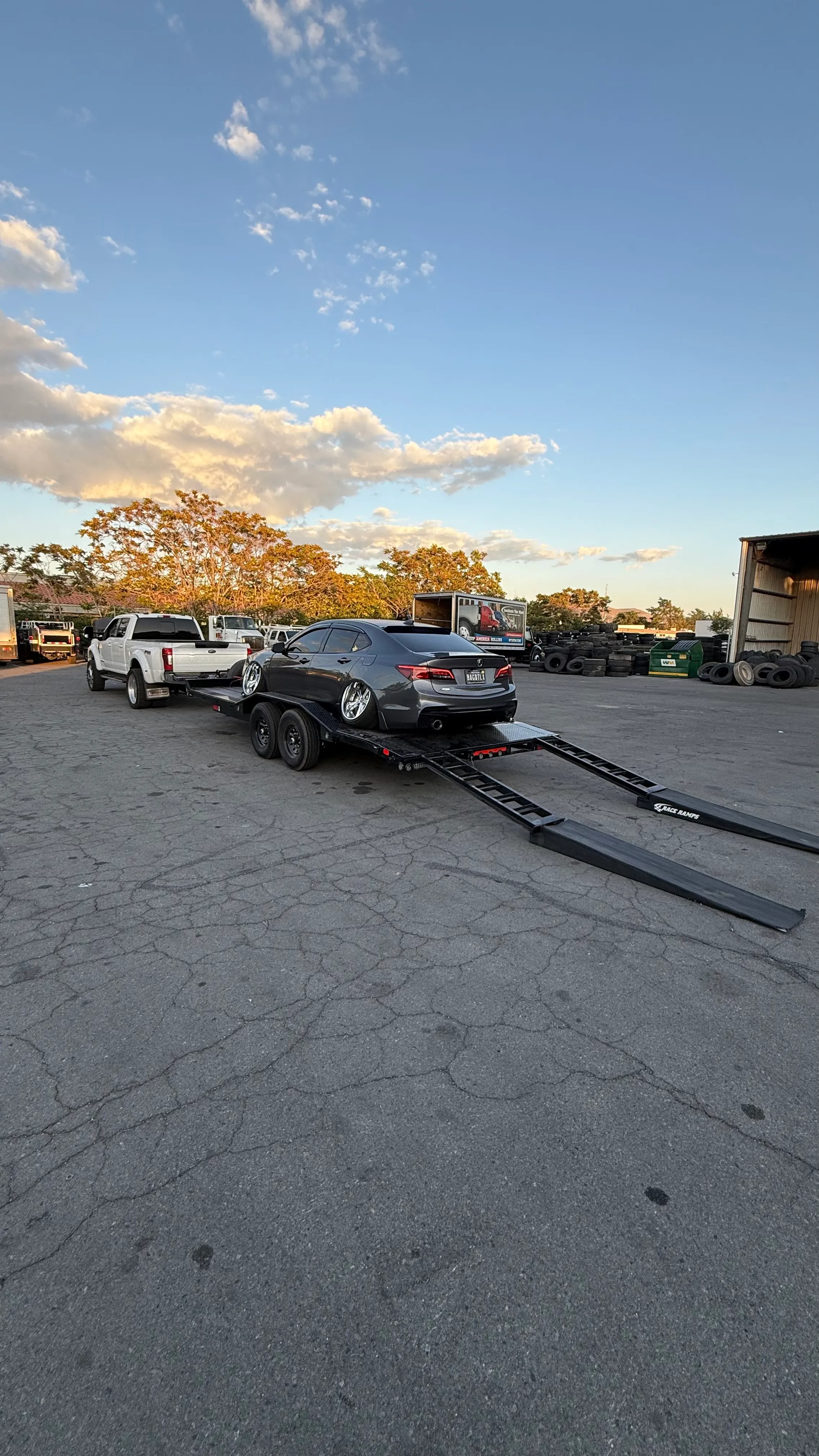 Dark gray sedan with lowered wheels on a car trailer attached to a white pickup truck in a parking lot.