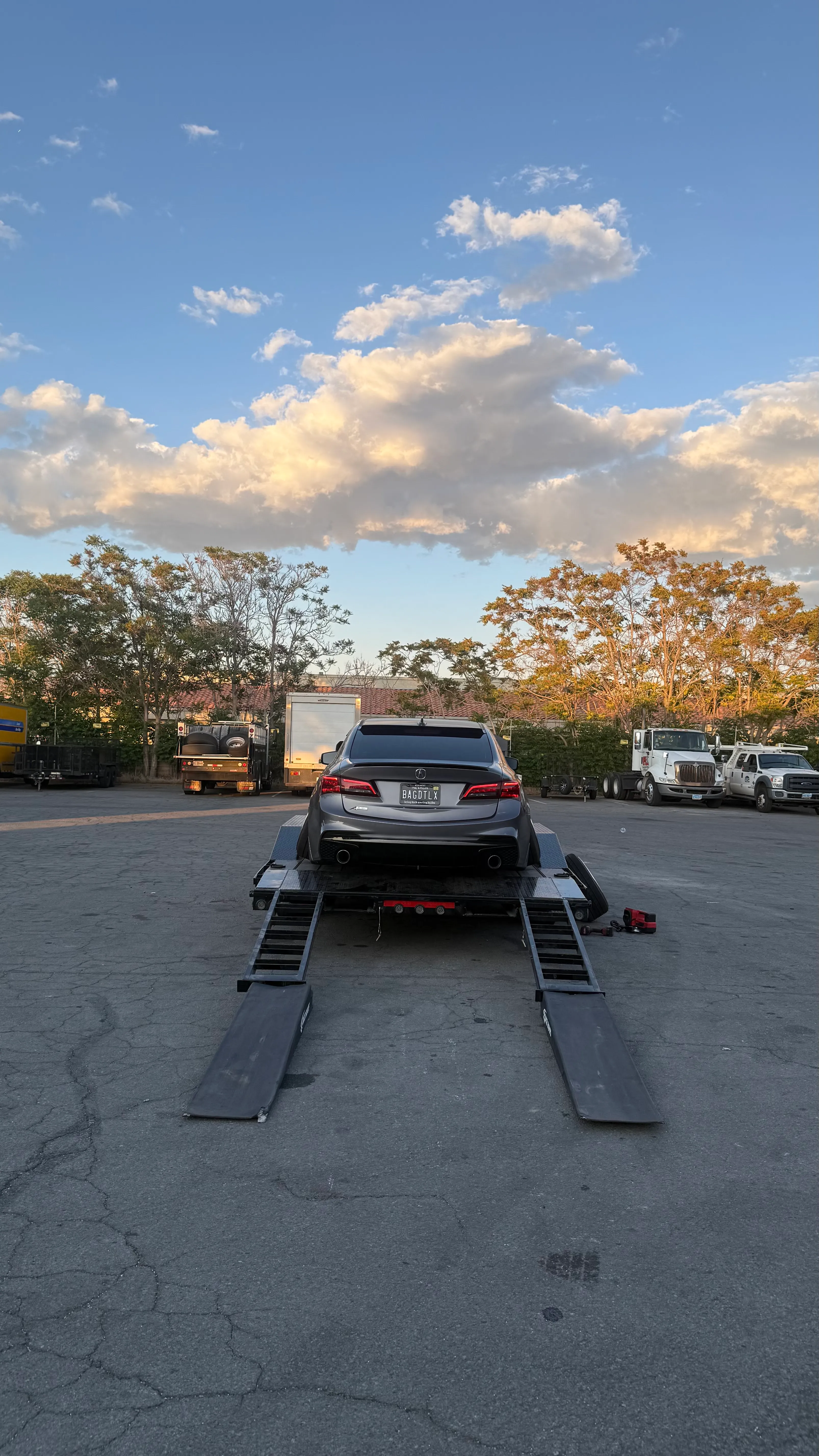 Black car with license plate BAGDTLX on a trailer ramp in a parking lot with trucks and trees.