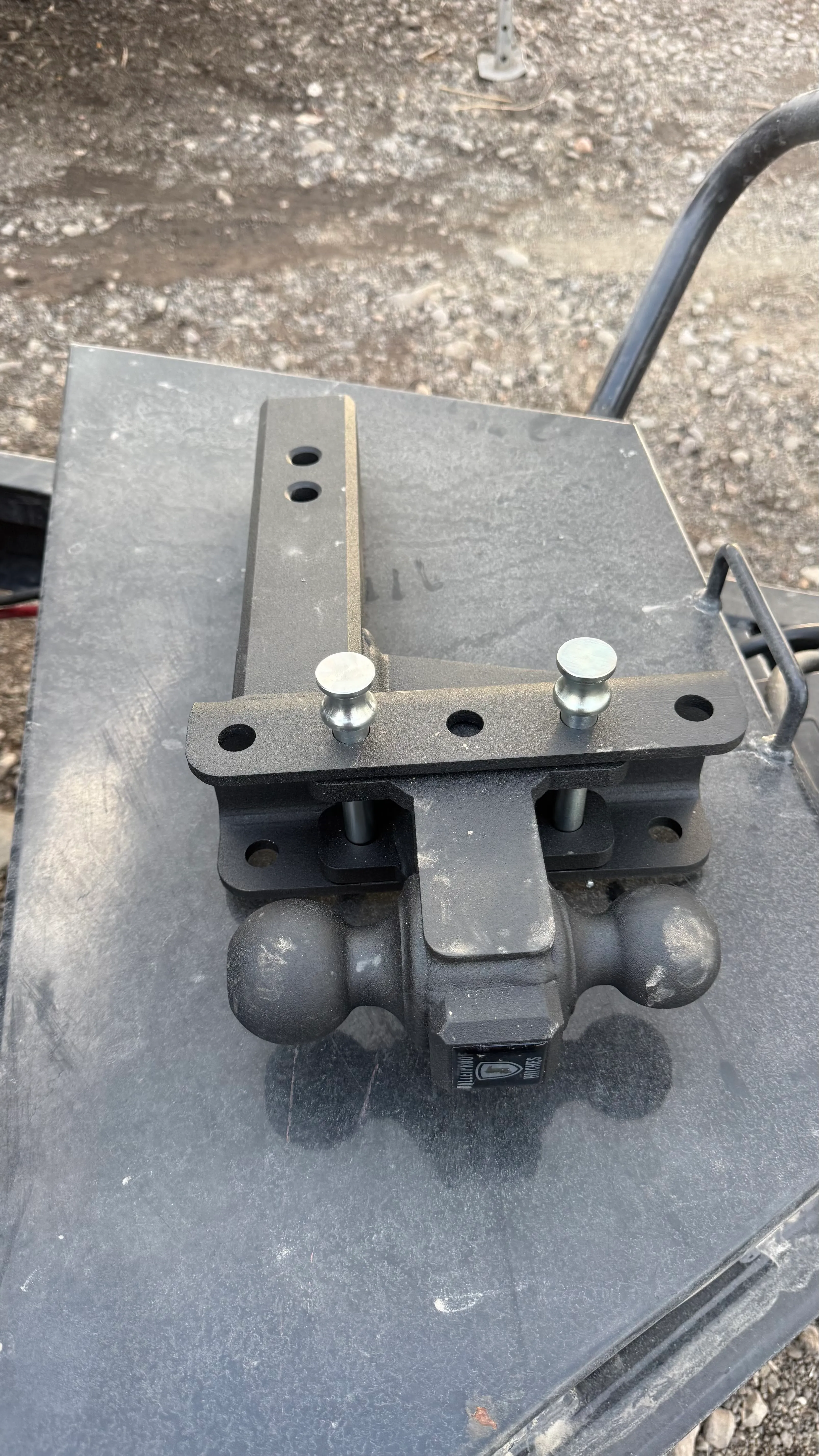 Black dual ball trailer hitch mounted on a flat metal surface outdoors on gravel ground.