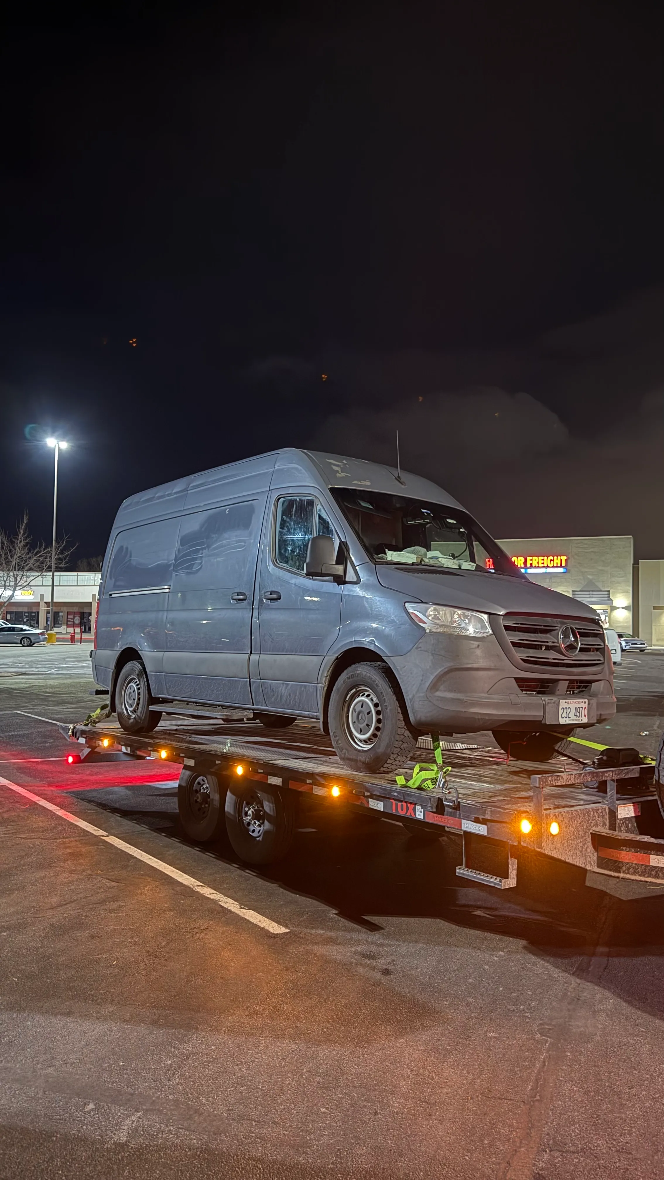 Gray Mercedes van secured on a flatbed trailer in a parking lot at night.