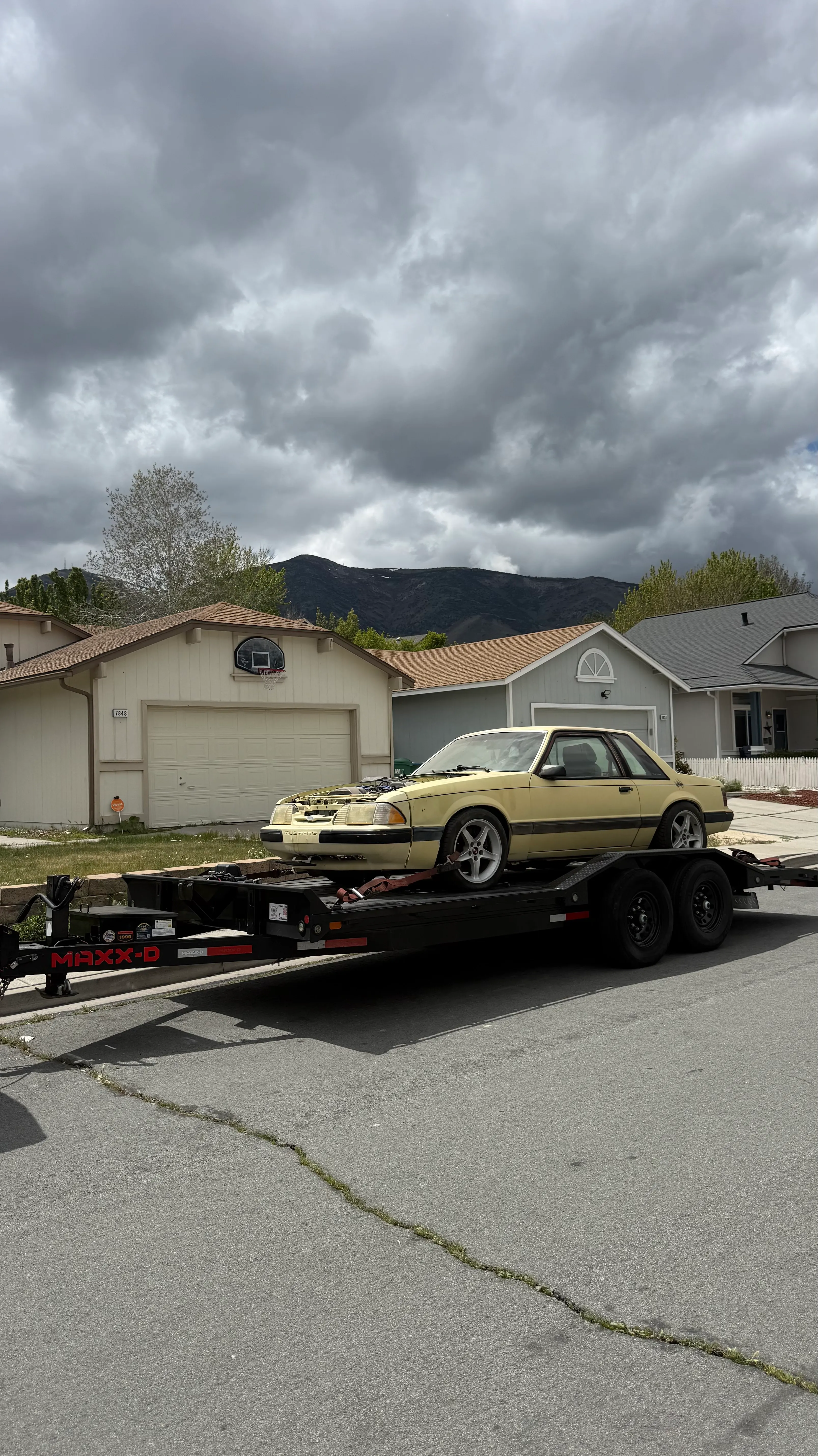 Yellow car without hood loaded on a black trailer parked on a suburban street with houses and mountains behind.