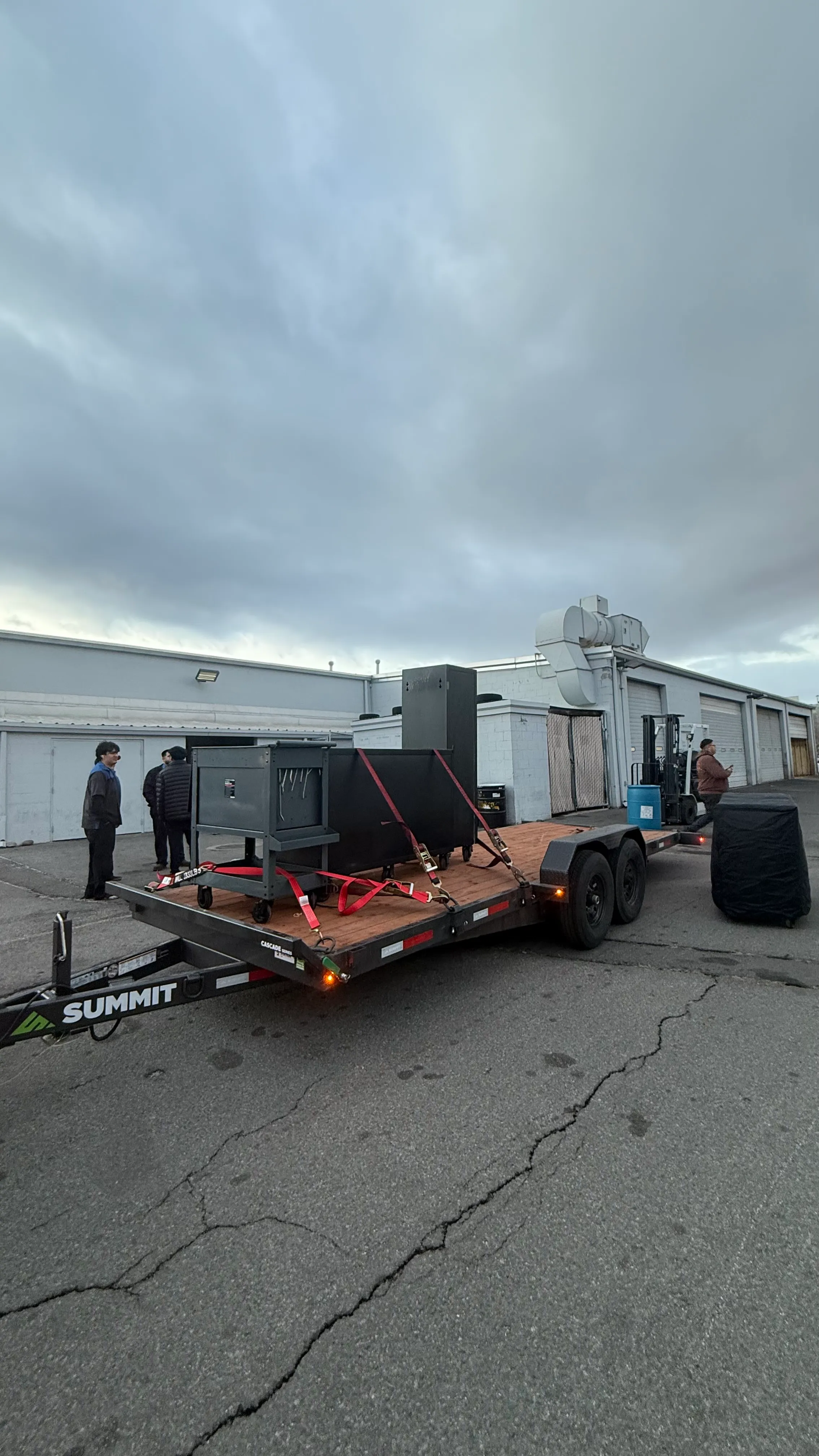 Two men near a trailer carrying black equipment secured with red straps in an industrial parking area.