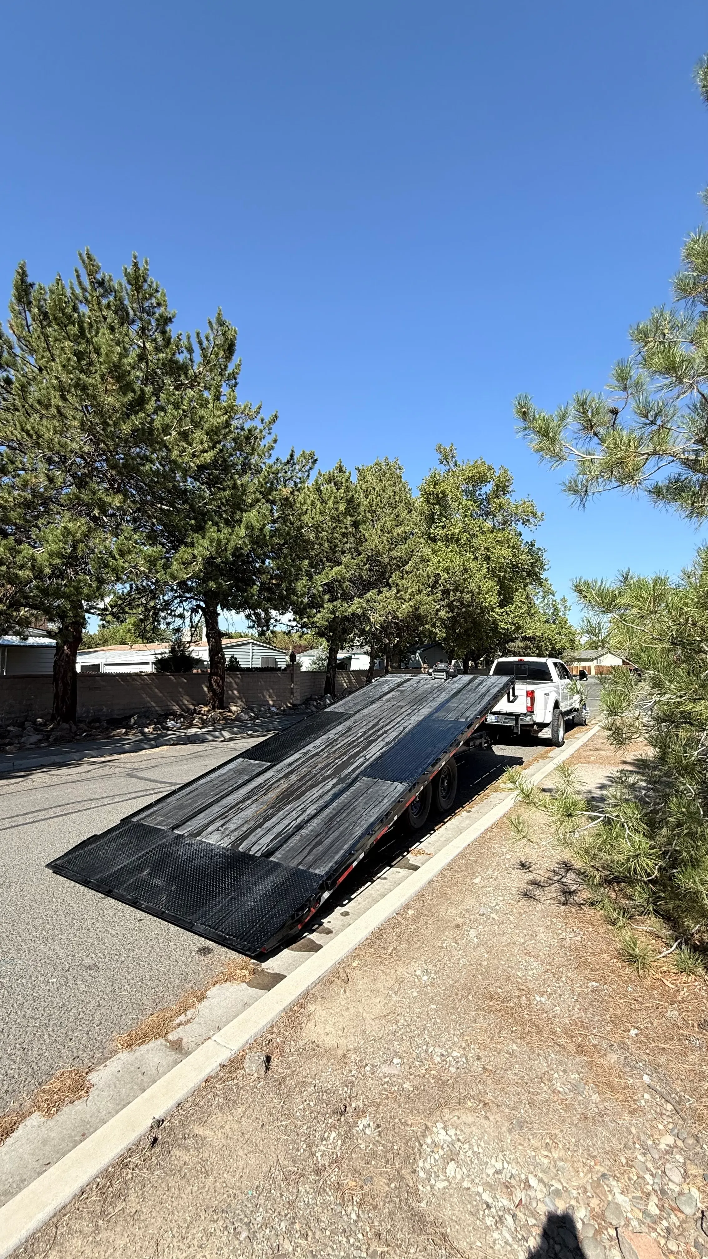 Tilted flatbed trailer hitched to white pickup truck parked on a sunny roadside with trees.