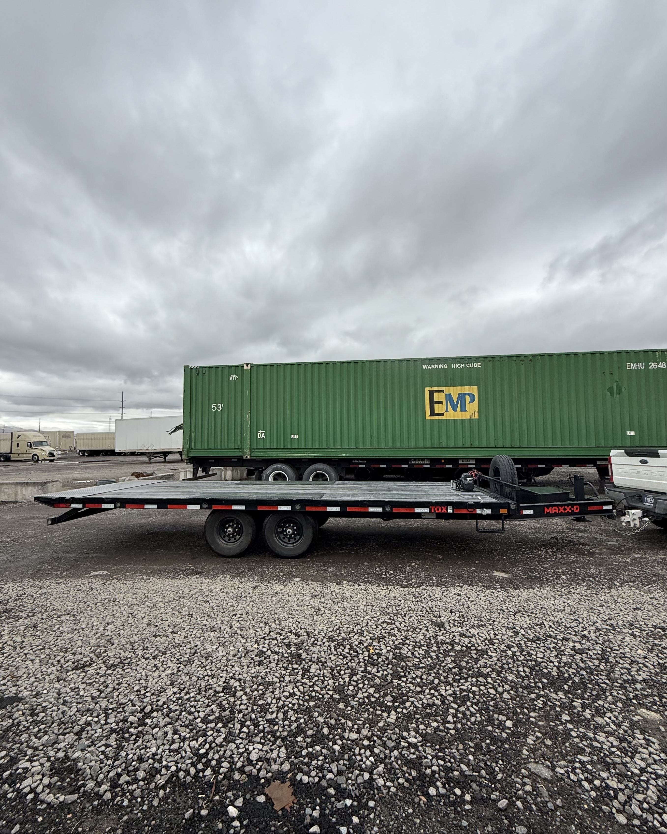 Black flatbed trailer with dual axles parked on gravel in front of a green EMP shipping container under cloudy sky.