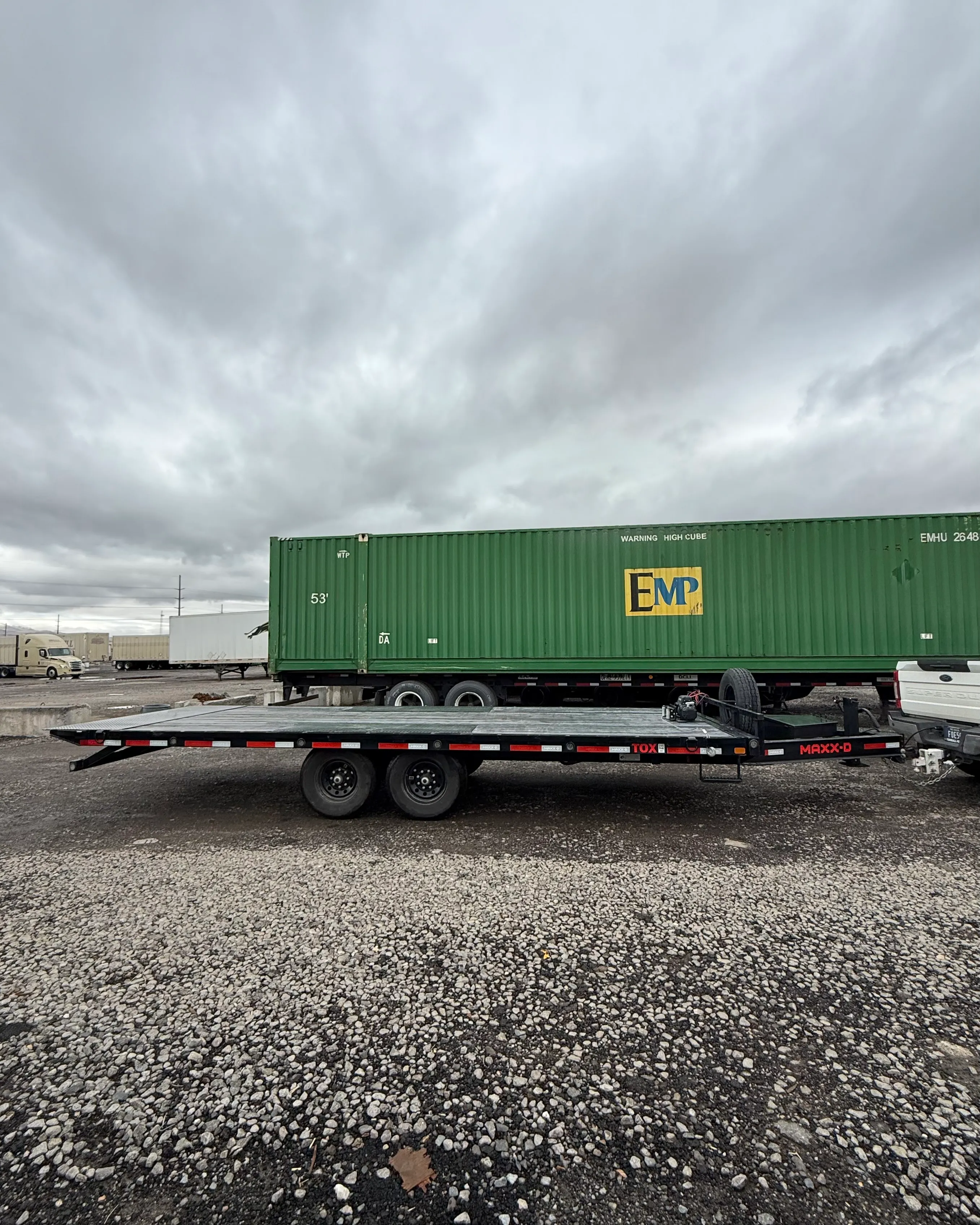 Flatbed trailer parked on gravel with a green EMP shipping container and cloudy sky in the background.