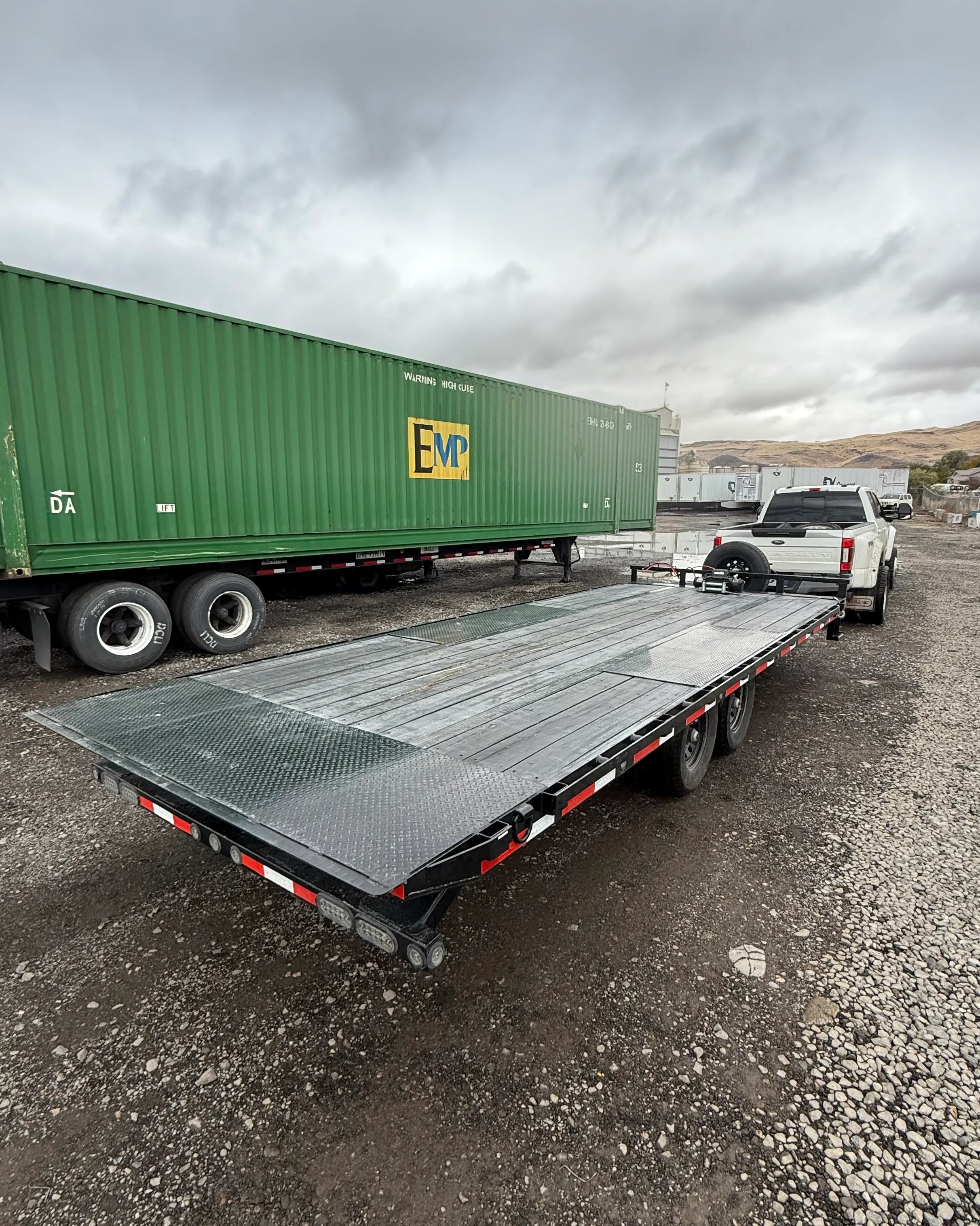 White pickup truck towing a flatbed trailer in a gravel lot with a green shipping container nearby.