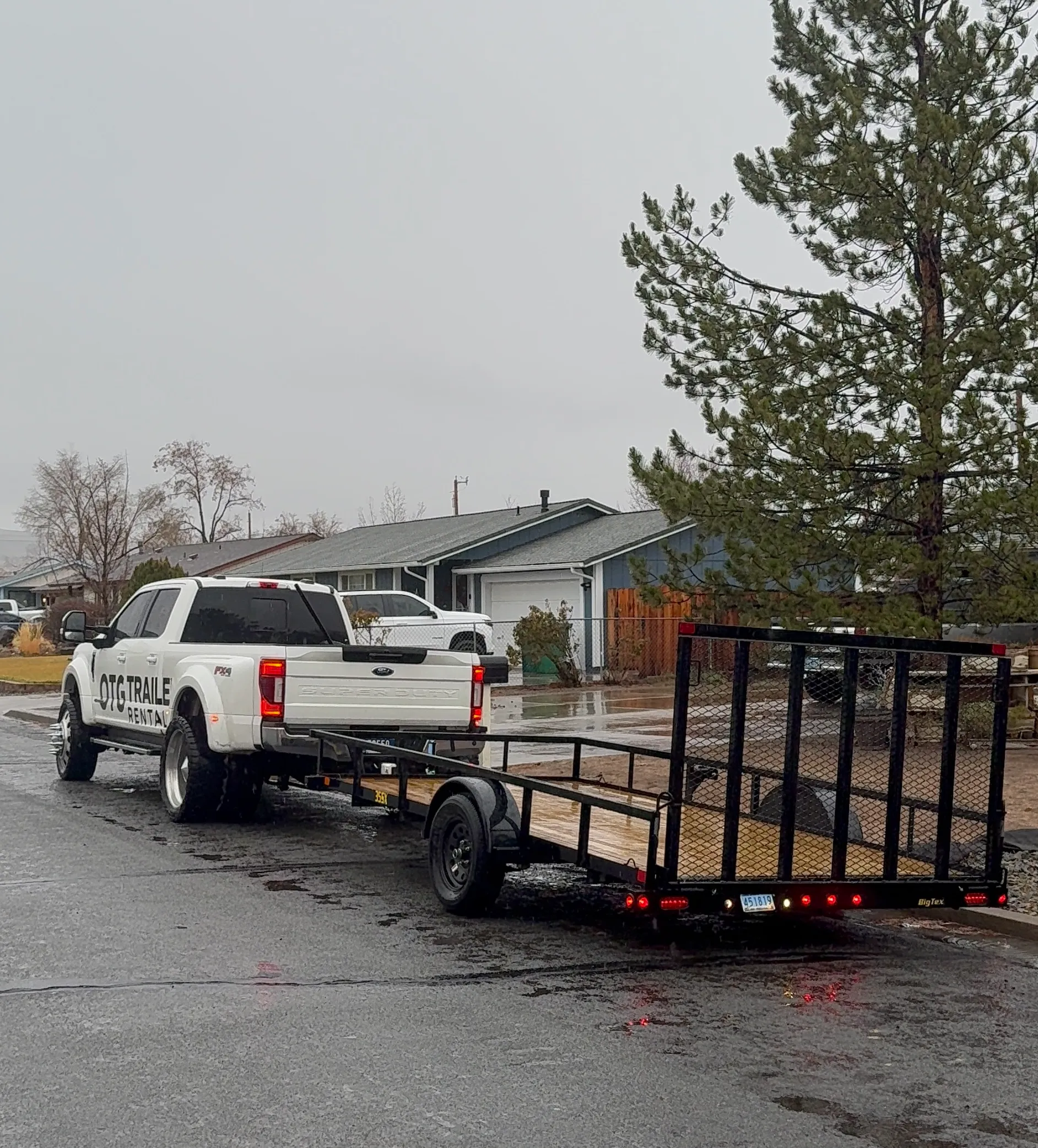 White pickup truck towing a flatbed trailer parked on a wet residential street on a cloudy day.