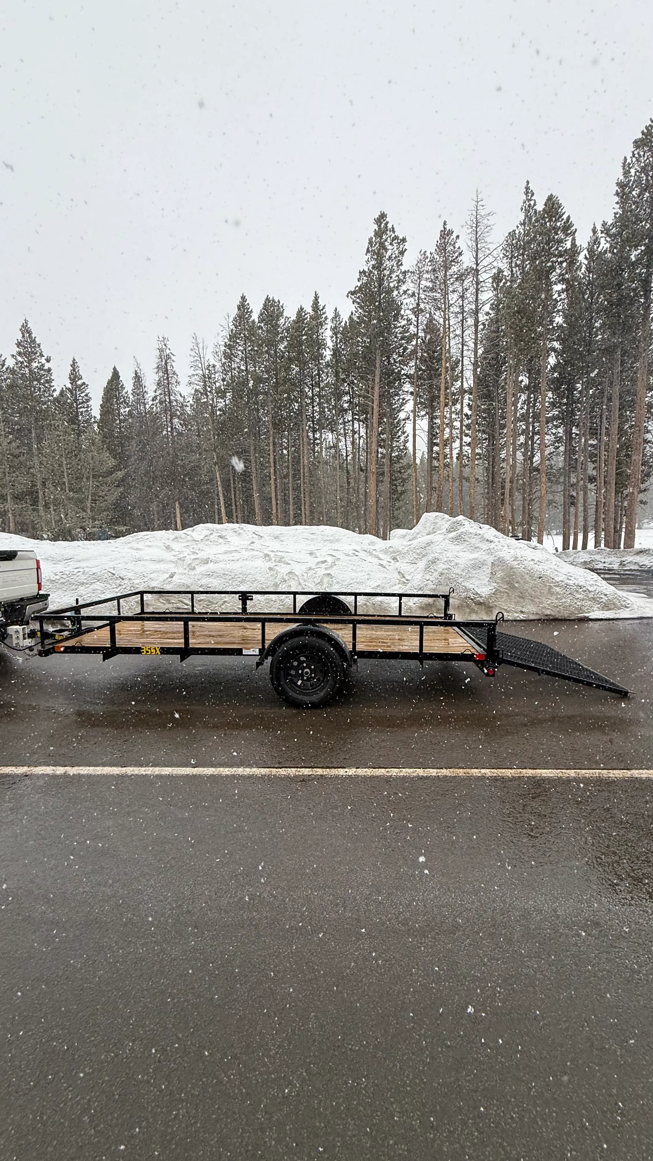 Flatbed utility trailer hitched to a white truck with snow and pine trees in the background.