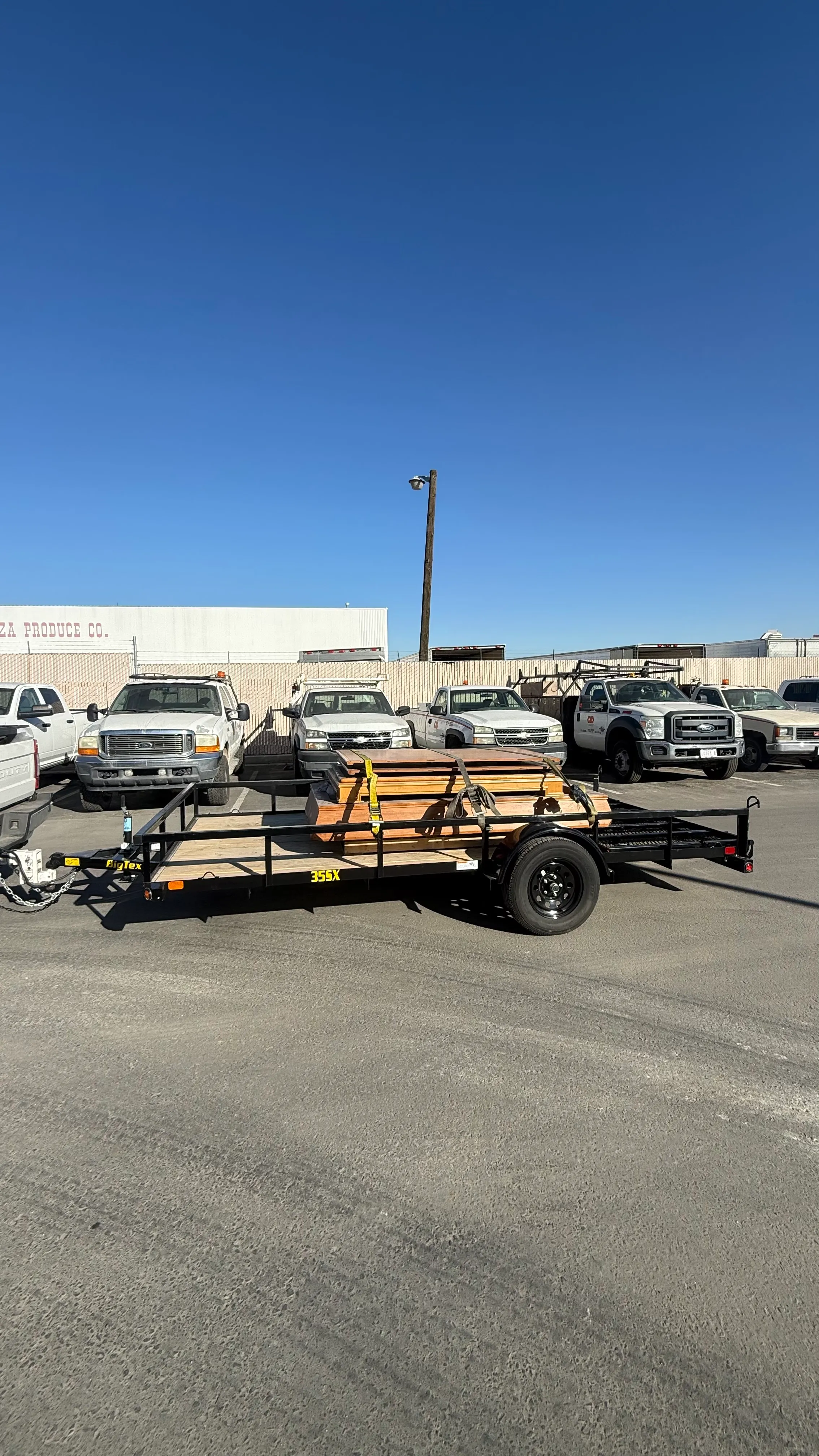 Flatbed trailer loaded with wooden planks parked in front of a row of pickup trucks under clear blue sky.