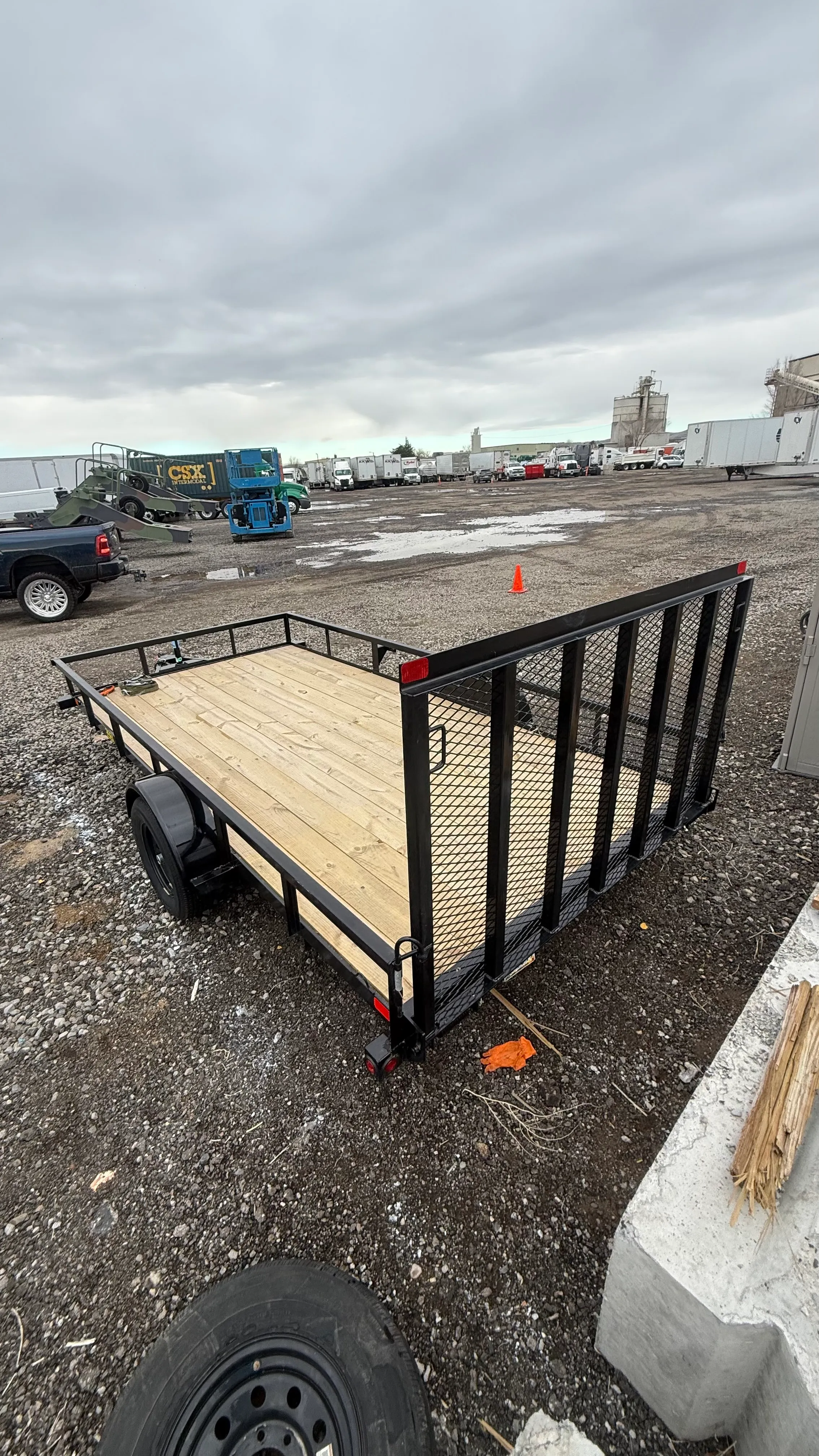 Empty utility trailer with wood floor and metal mesh ramp in a gravel lot under cloudy sky.