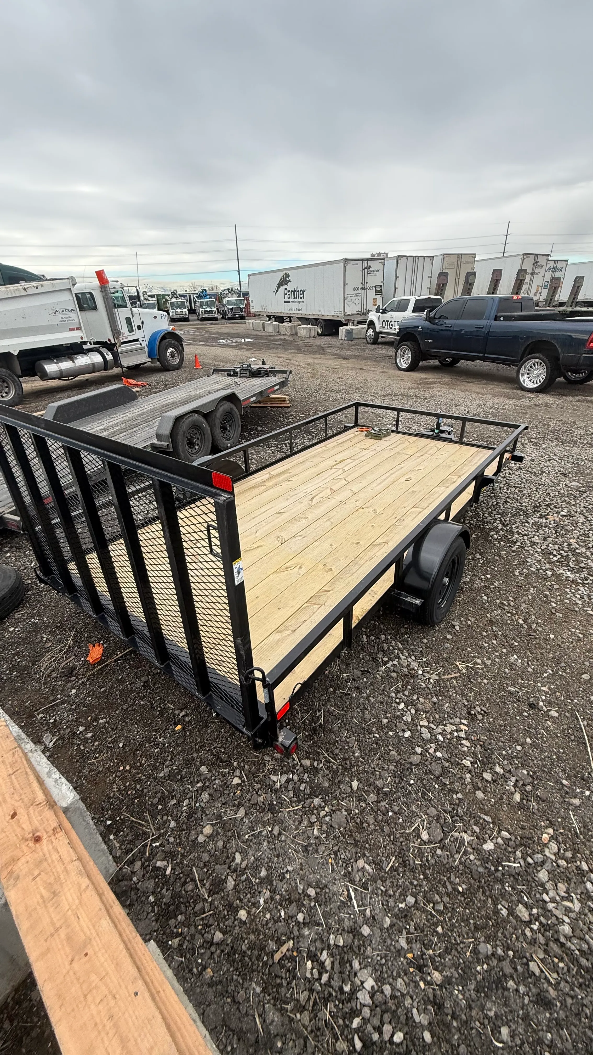 Empty small utility trailer with wooden floor and metal frame parked on gravel in a truck yard.